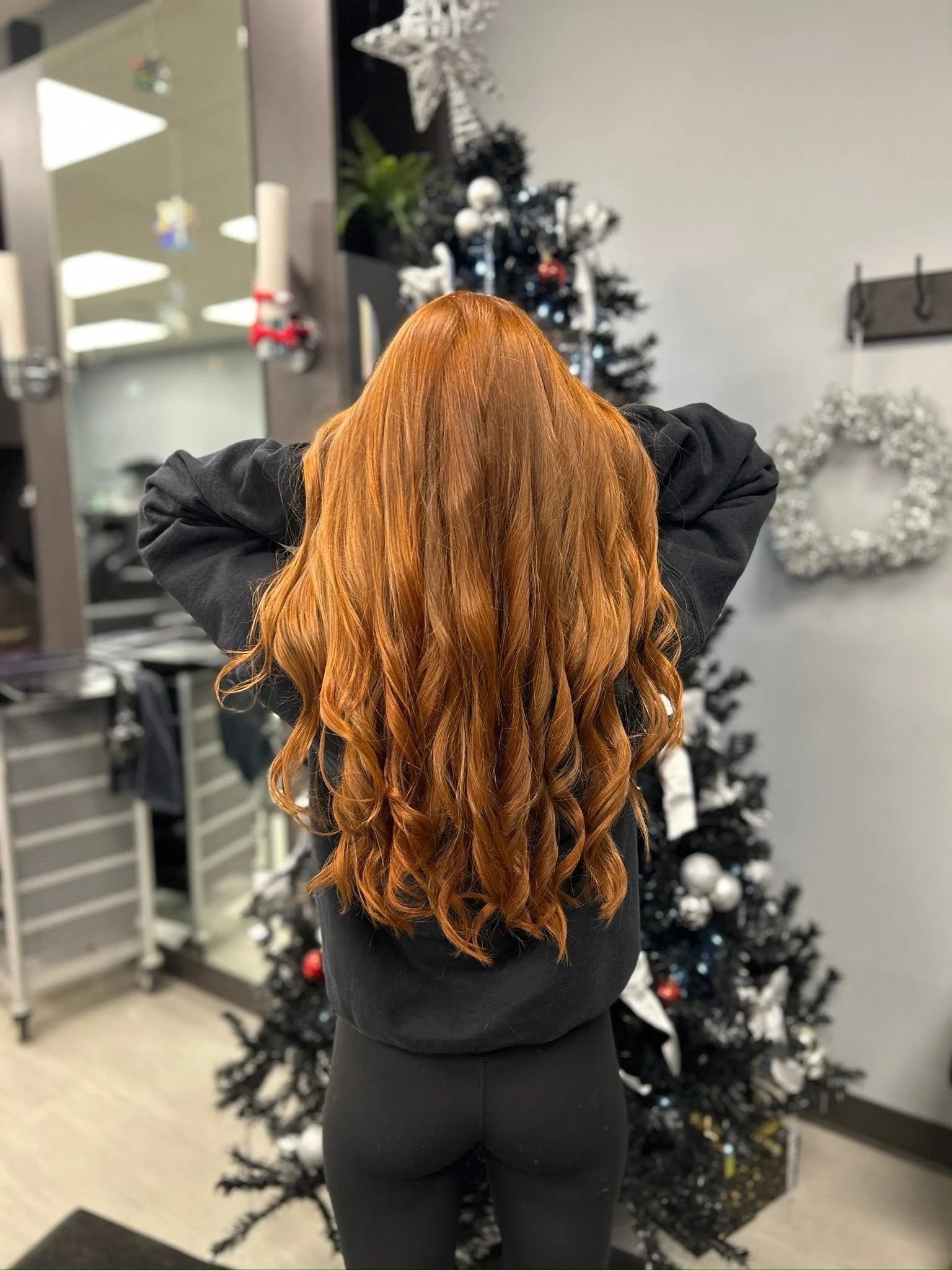 Back view of a woman with long, wavy, red hair standing in front of a decorated Christmas tree in a salon.