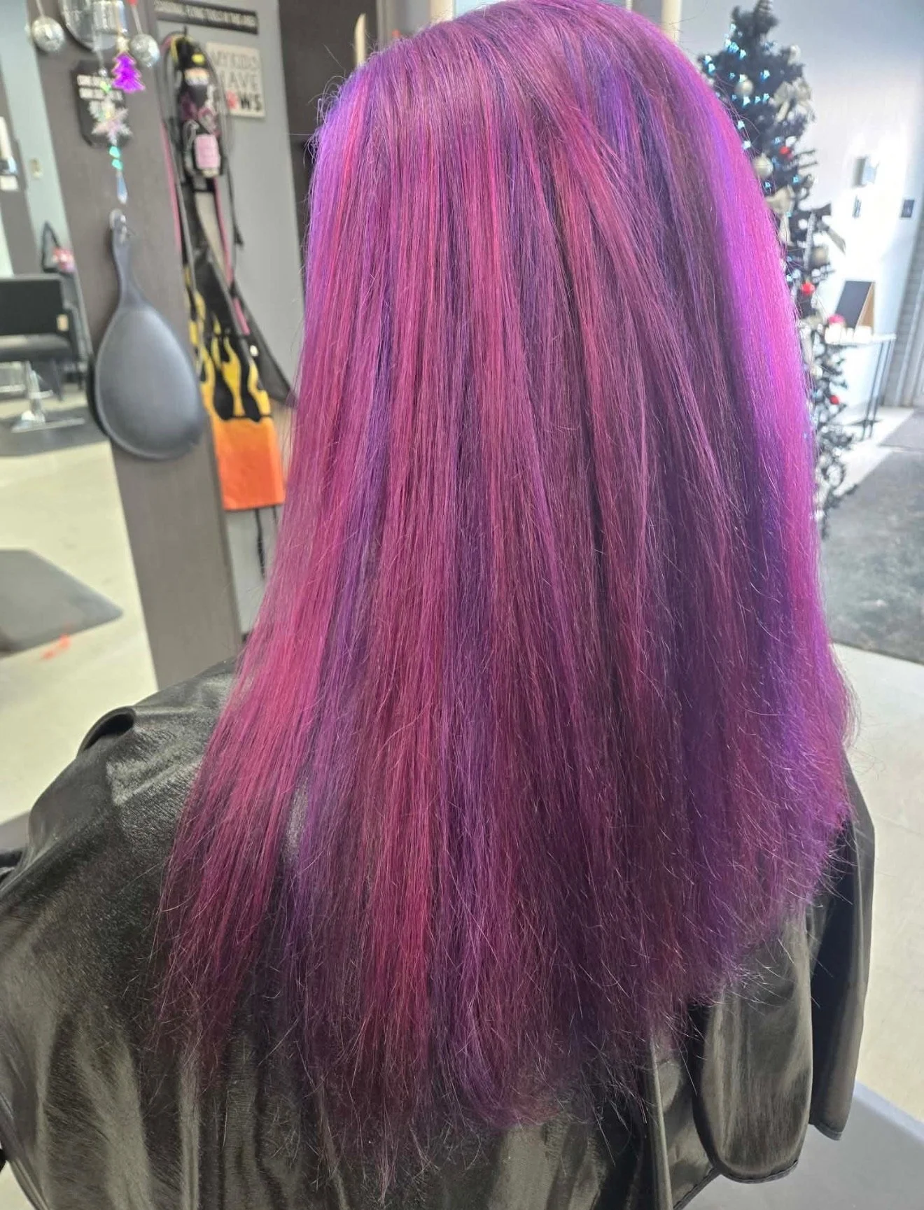 Back view of a woman with straight, vibrant, purple and pink ombre hair, standing in a salon with a Christmas tree in the background.