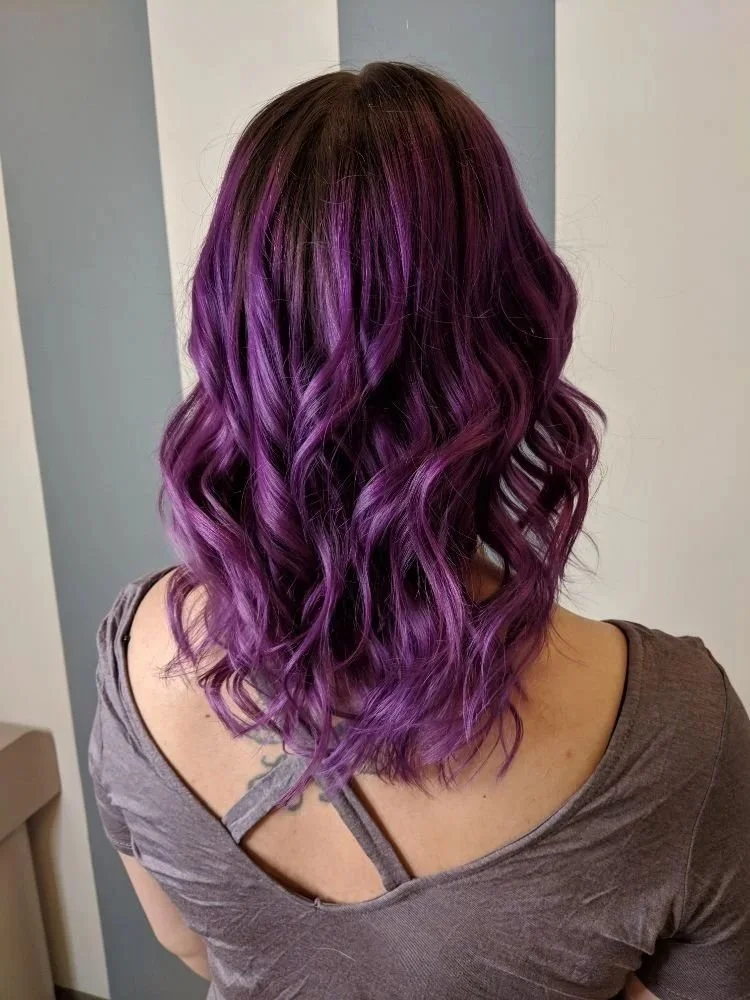 A woman with shoulder-length hair styled in loose waves, dyed in a gradient of dark purple at the roots to vibrant purple at the tips.