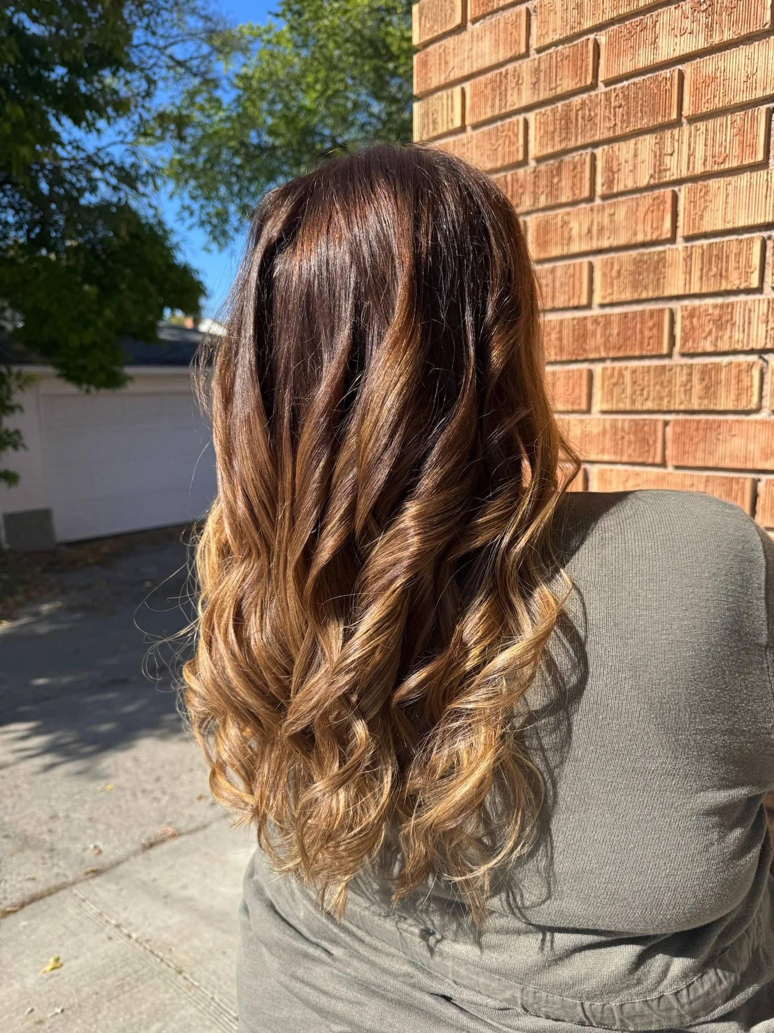 A person sitting outdoors with their back facing the camera, showing long, wavy brown hair with lighter highlights, against a brick wall, trees, and a bright blue sky in the background.
