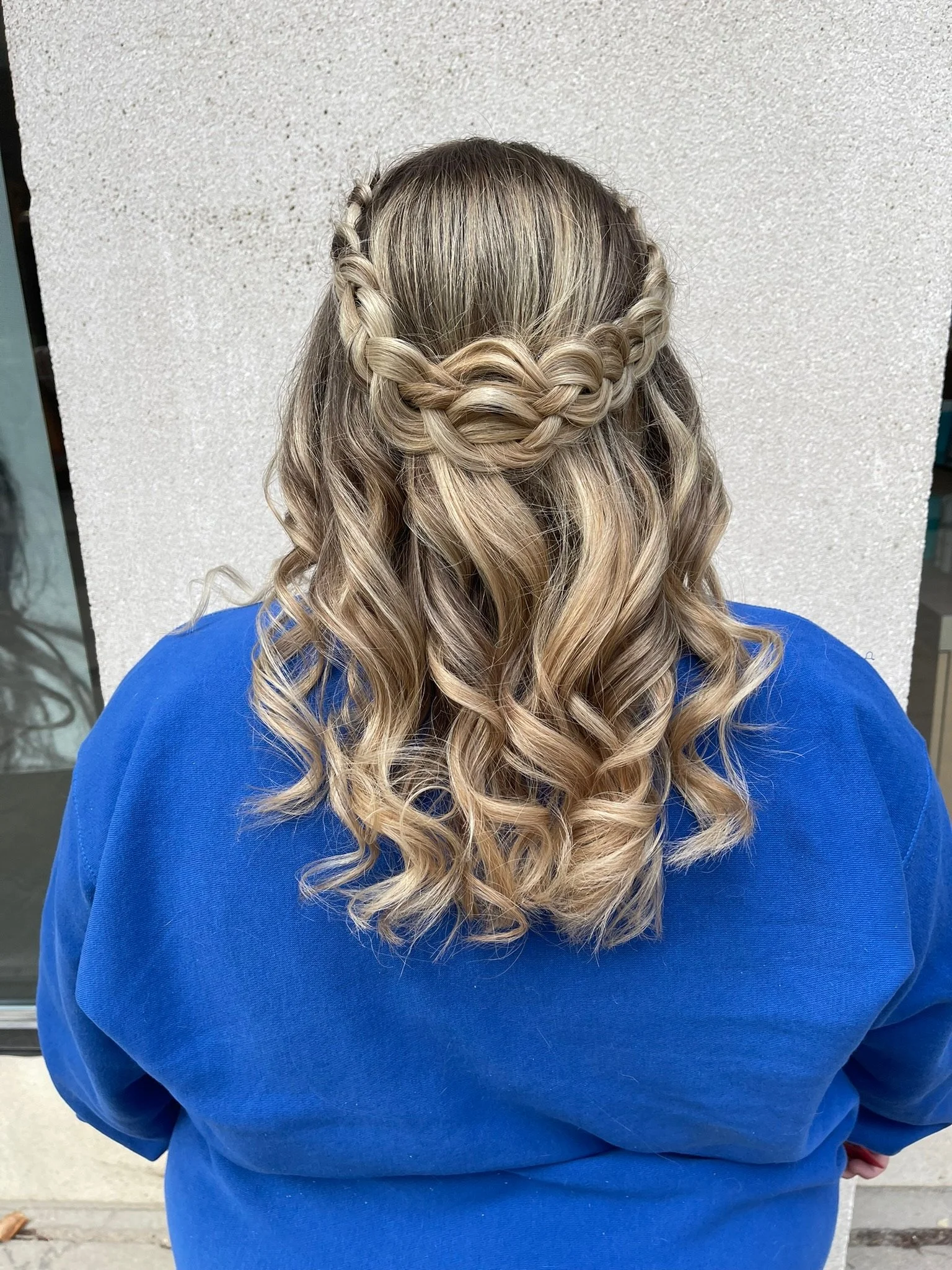 Back of a woman with blonde, curly hair styled with a braided crown, wearing a blue top, standing against a light-colored textured wall.