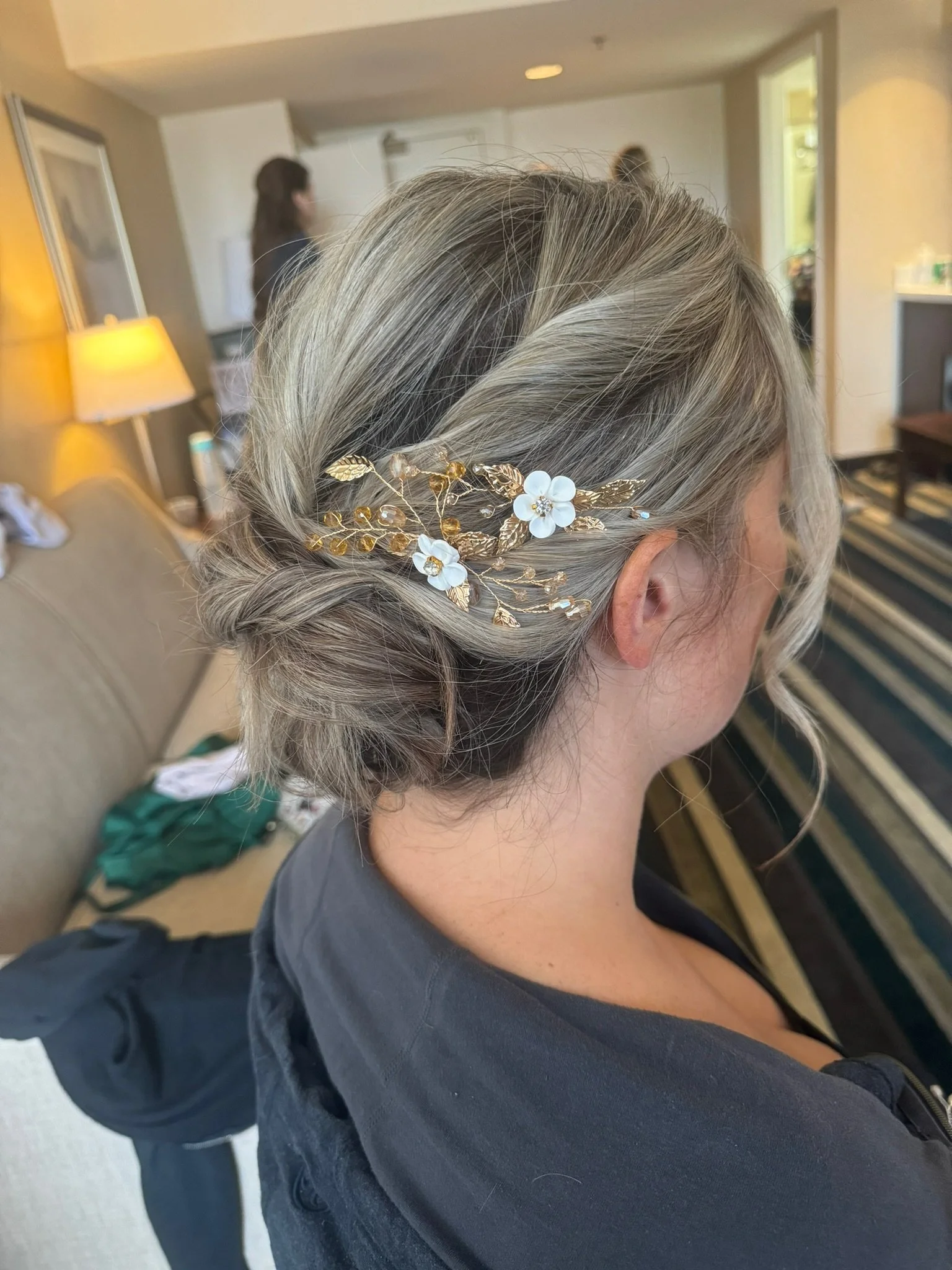 A woman with blonde hair styled in an elegant updo, decorated with a gold hairpiece featuring white flowers and leaves, indoors with a beige couch and a striped rug in the background.
