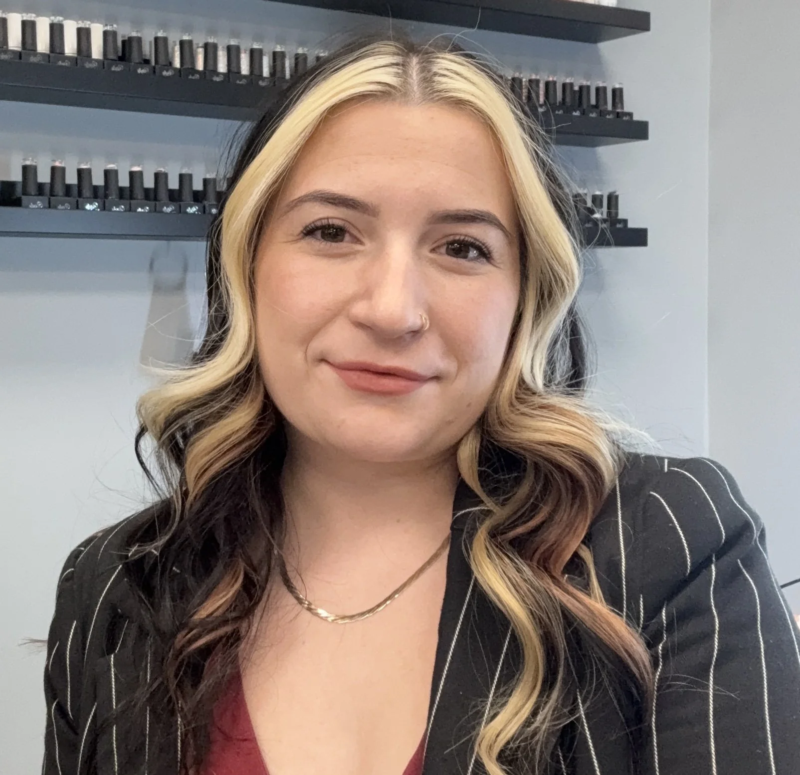 A young woman with blonde and brown hair in loose waves, wearing a black pinstripe blazer over a maroon top, smiling at the camera in a salon with shelves of nail polish in the background.