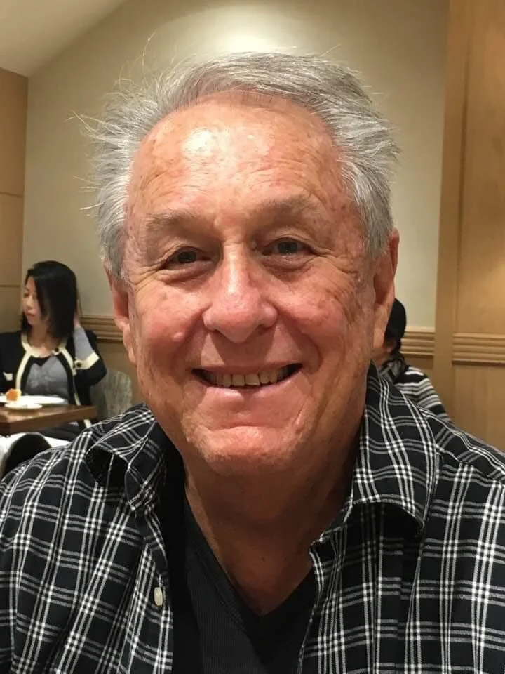 Older man with white hair smiling, wearing a black and white plaid shirt, in a restaurant