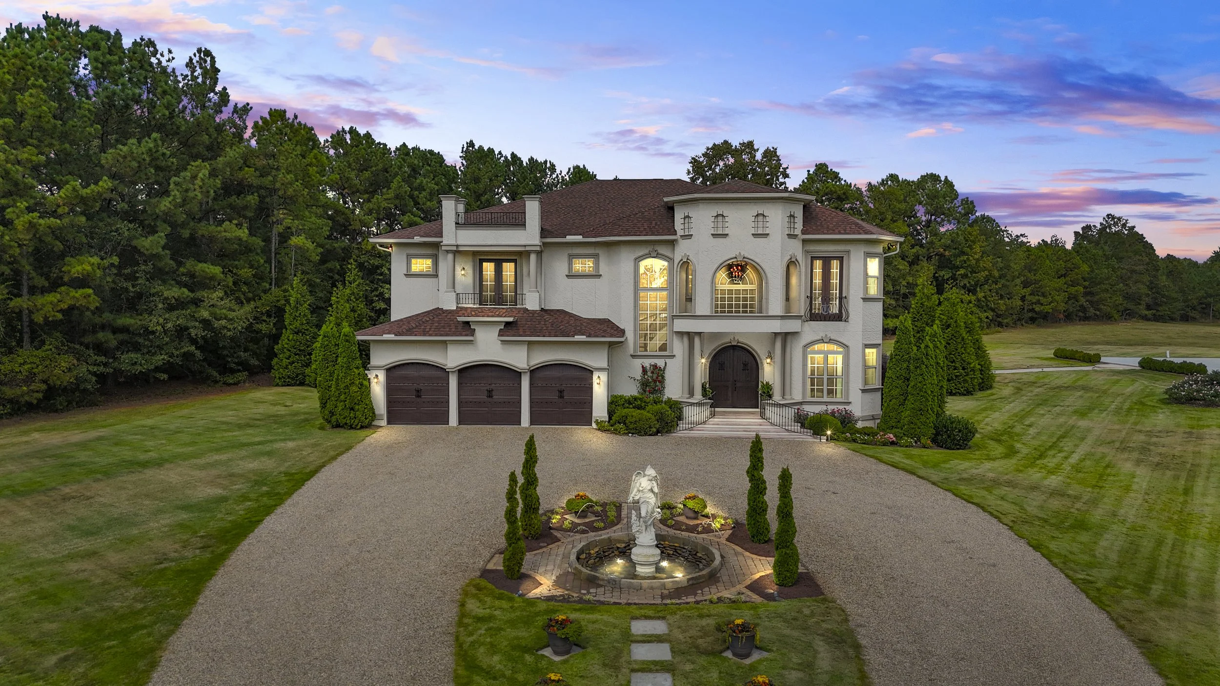 Large estate home with three-car garage, well-manicured lawn, and fountain in front, surrounded by trees at sunset.