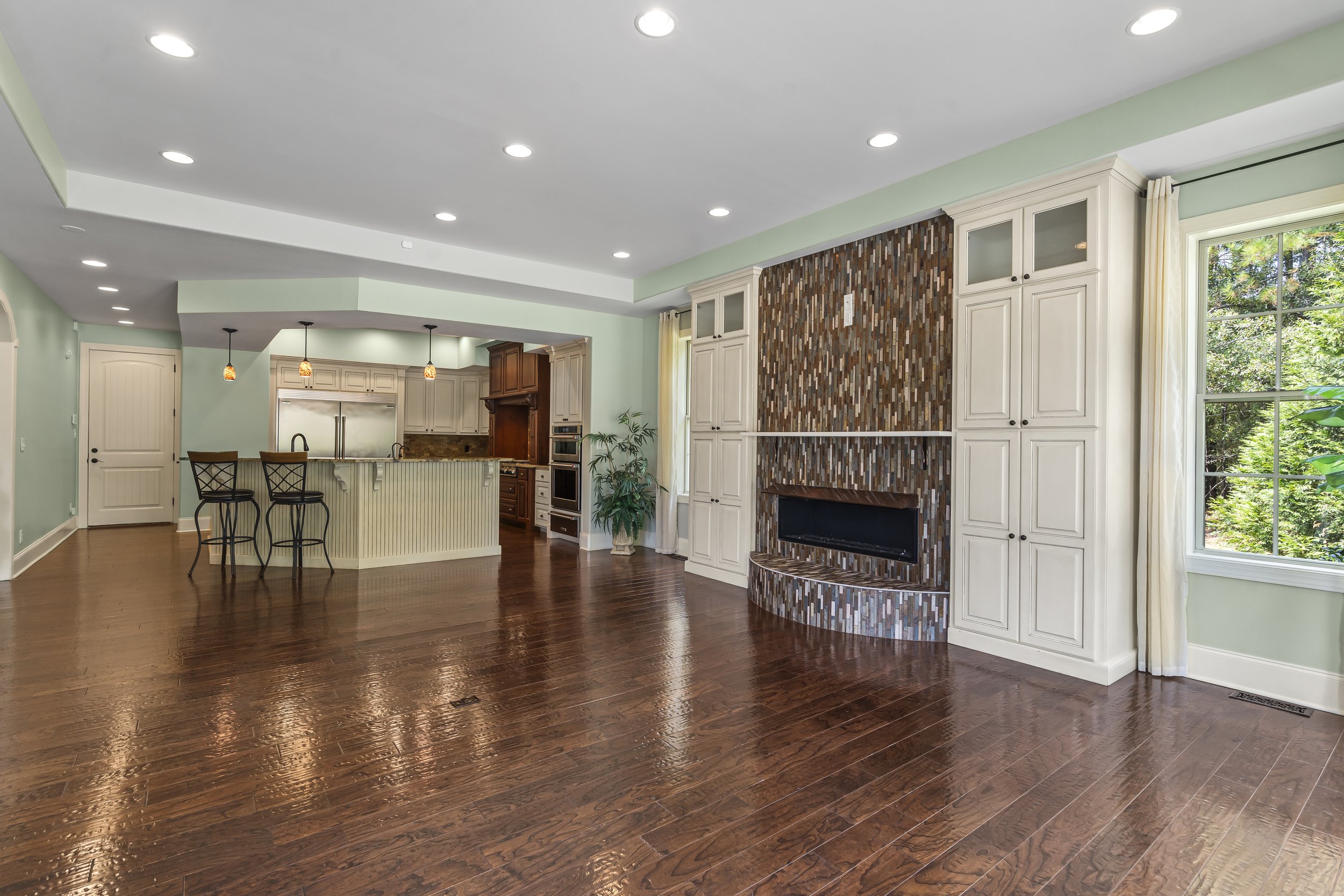 Spacious living room with hardwood floors, a wood-paneled fireplace, white built-in cabinetry, large windows, and an open kitchen with a breakfast bar.