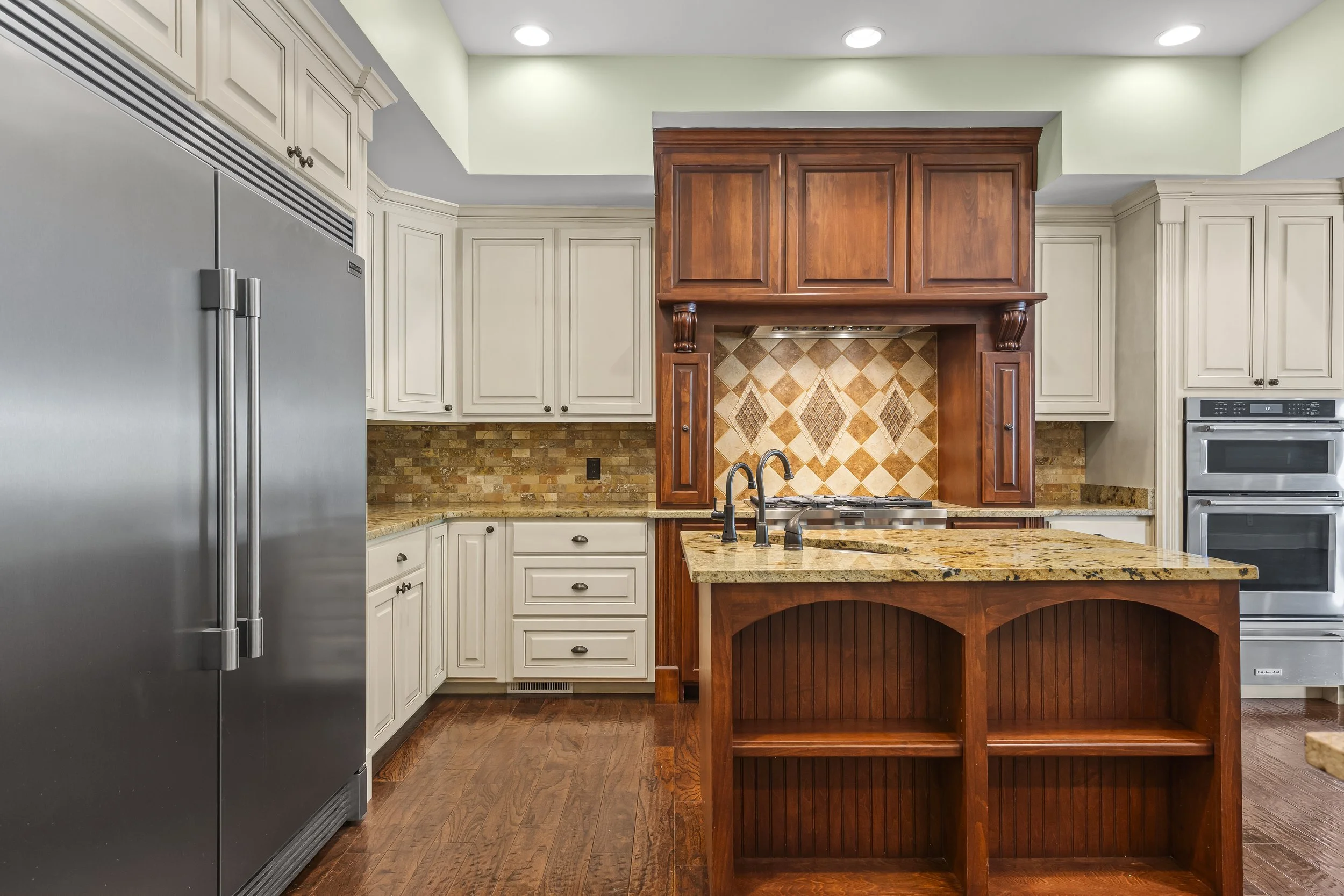 A kitchen with white cabinets, a stainless steel refrigerator, a wooden island with a granite countertop, and a tiled backsplash.