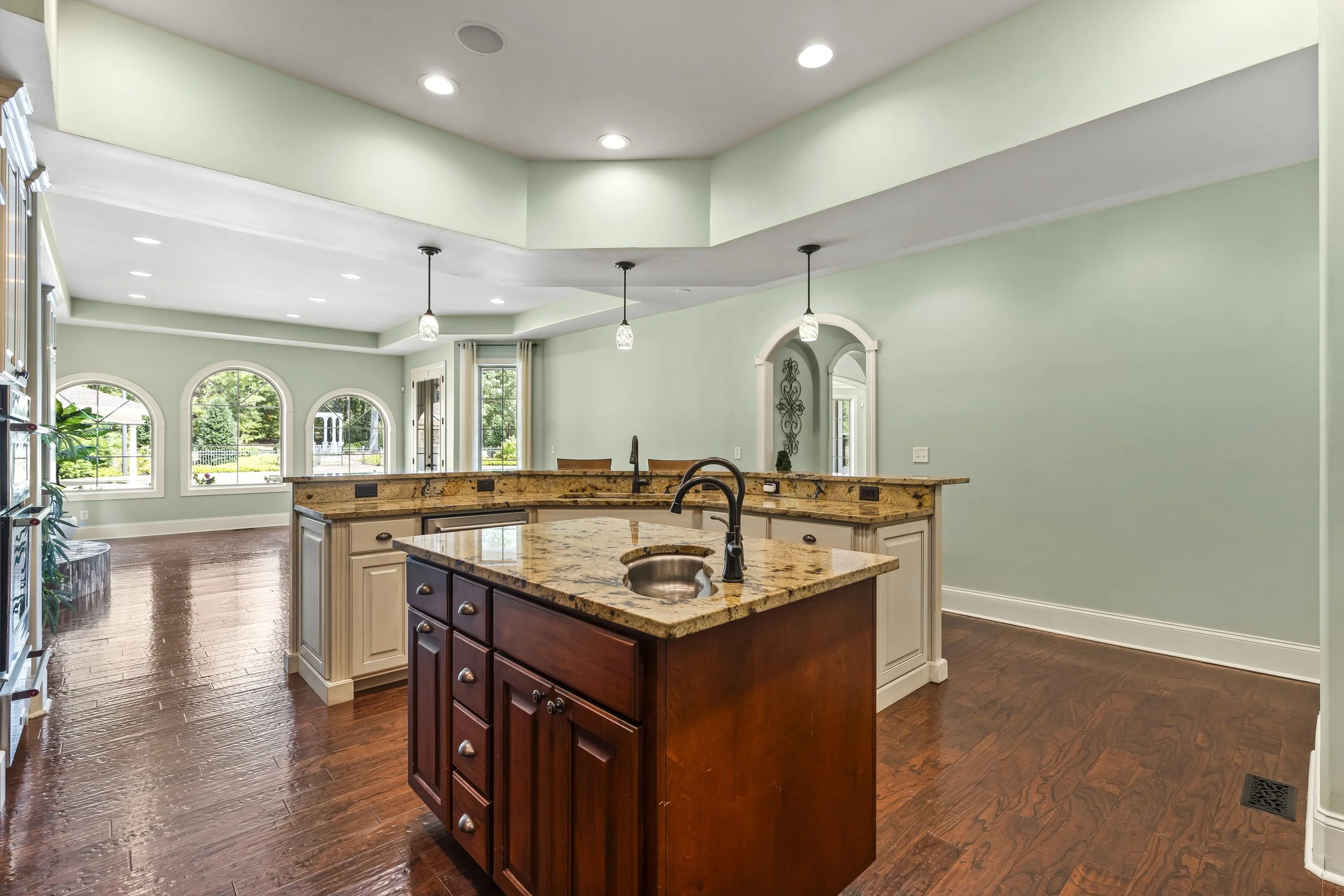 Open kitchen with wooden floors, green walls, a kitchen island with a granite countertop, and large arched windows in the background.
