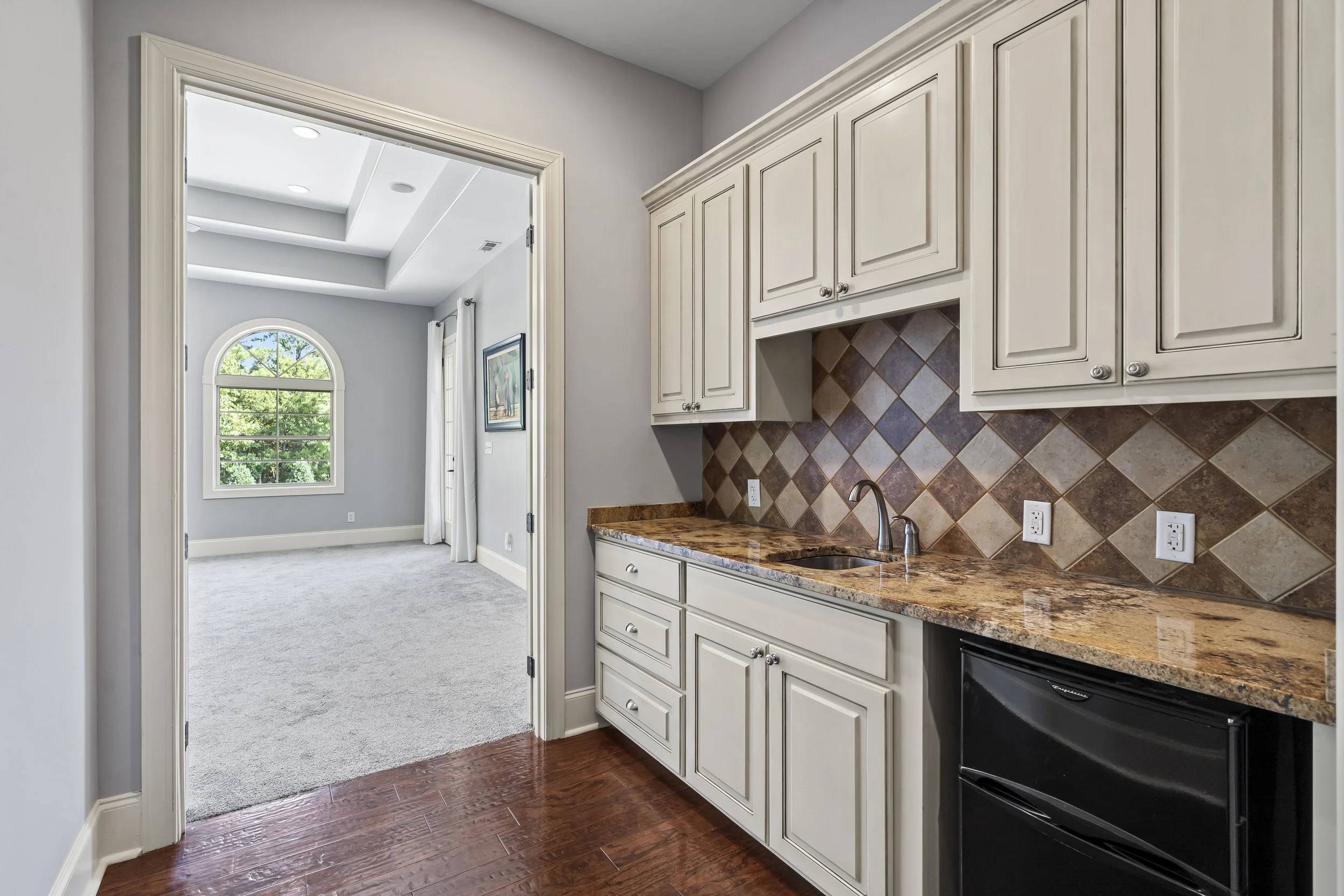 Kitchen area with cream cabinets, a granite countertop, and a tiled backsplash, with a view into a bright living room with a large arched window.