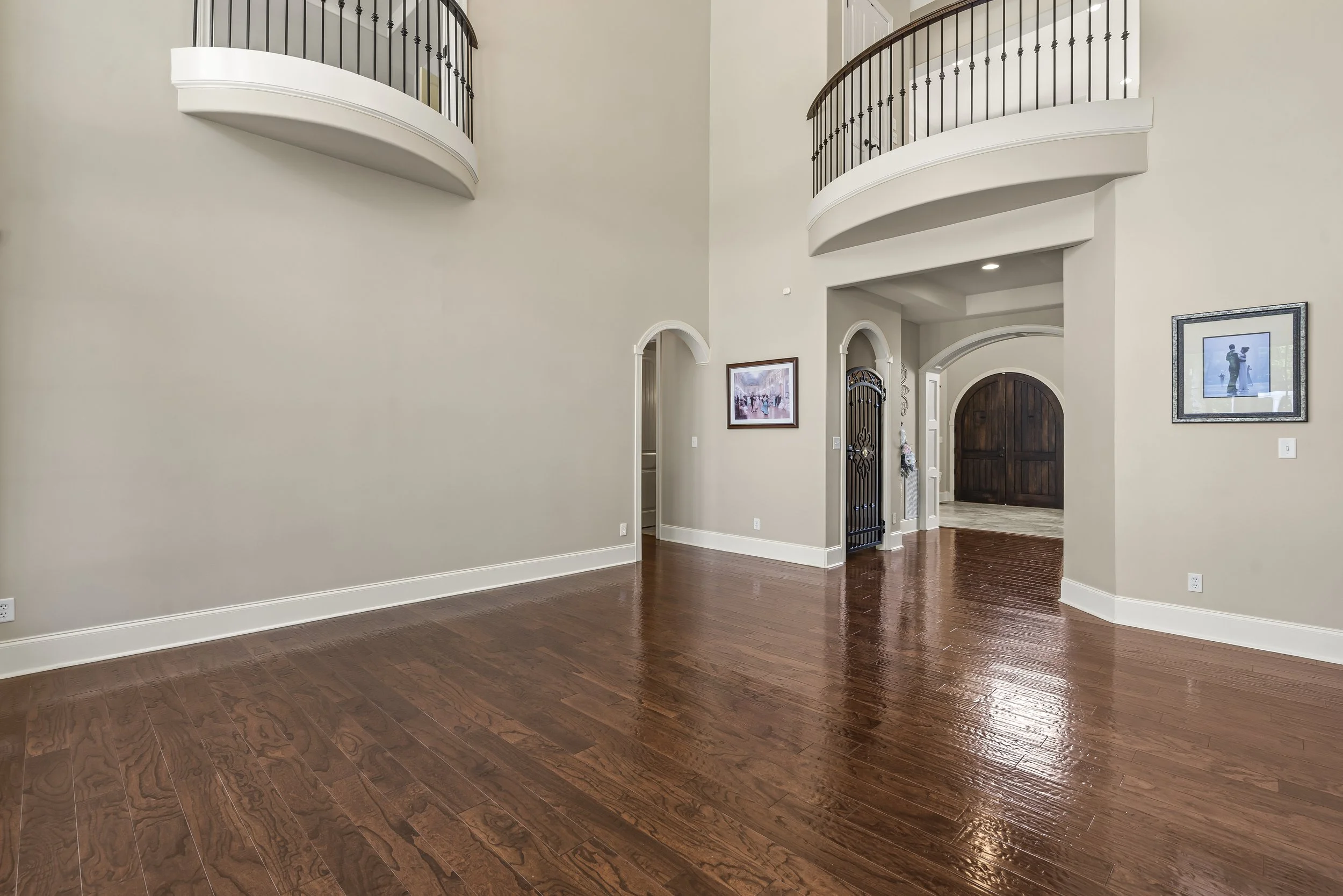 Empty living room with polished hardwood floors, beige walls, two upper balconies with black railings, and a dark wooden front door.