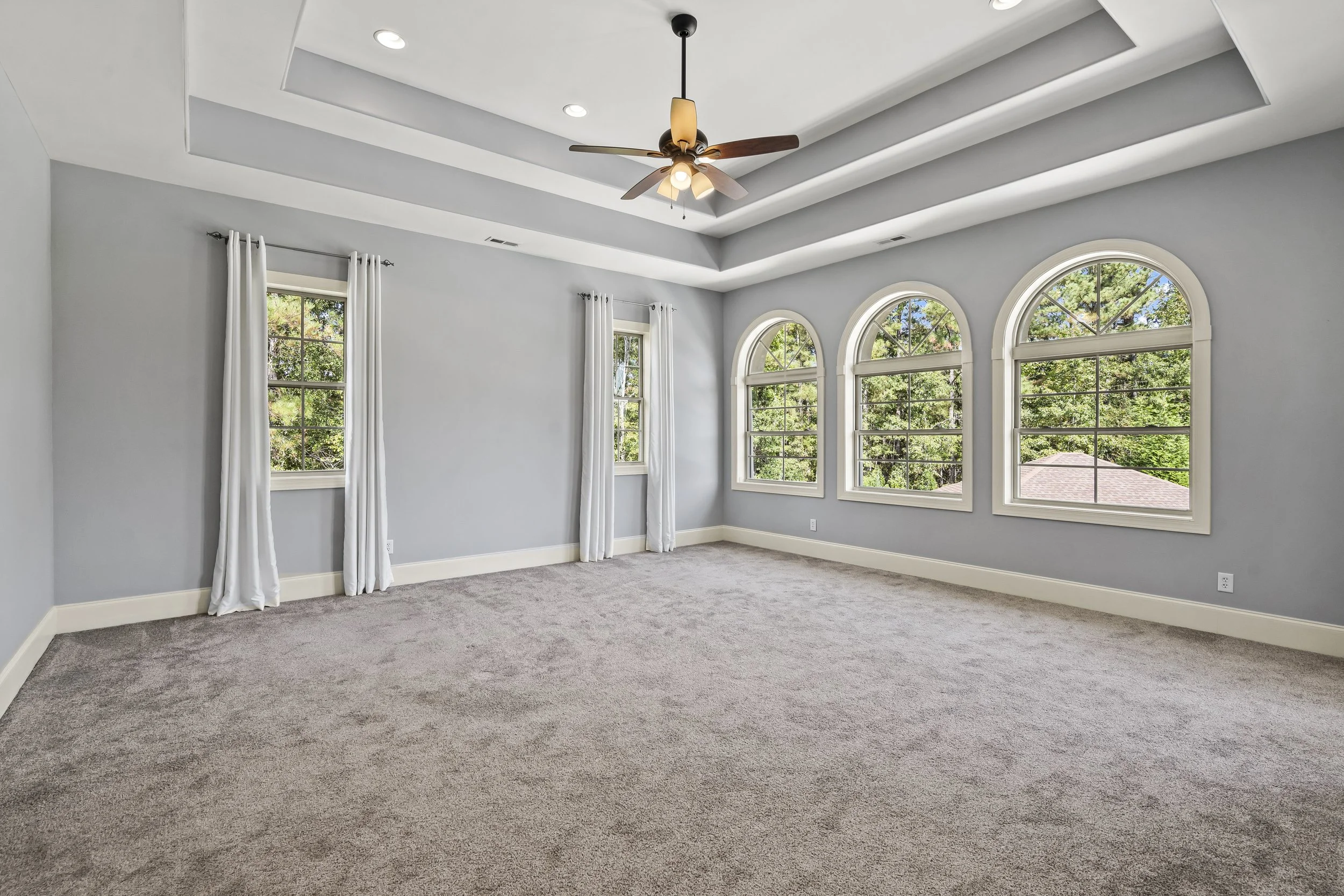 Empty bedroom with large arched windows, white curtains, carpeted floor, and a ceiling fan with lights.