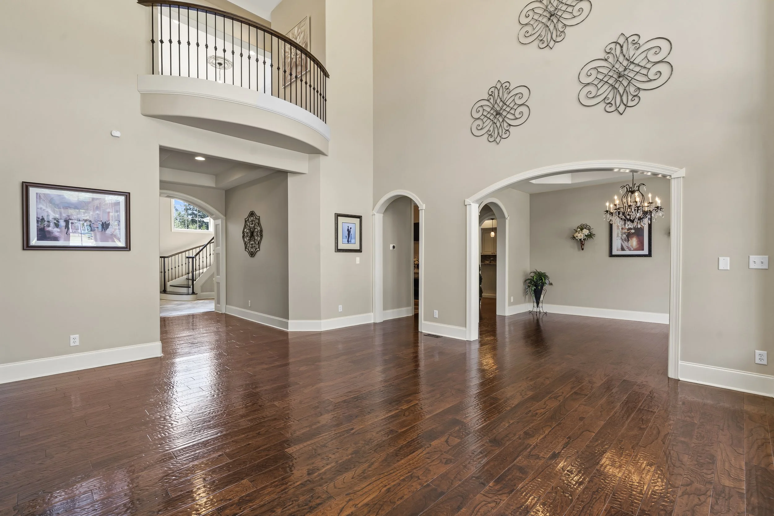 Interior view of a spacious living area with wooden floors, arched doorways, wall art, decorative wall accents, and a chandelier. There is a staircase with black railing leading to an upper level.
