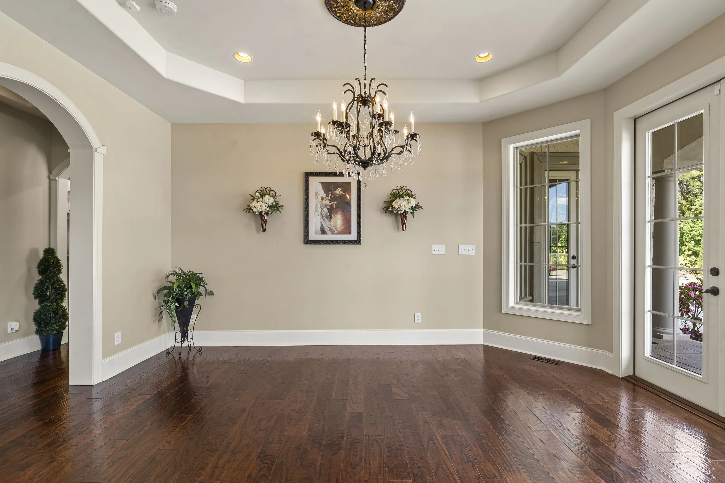 Empty living room with wooden floors, a chandelier hanging from the ceiling, wall decor, and large windows with a door leading outside.