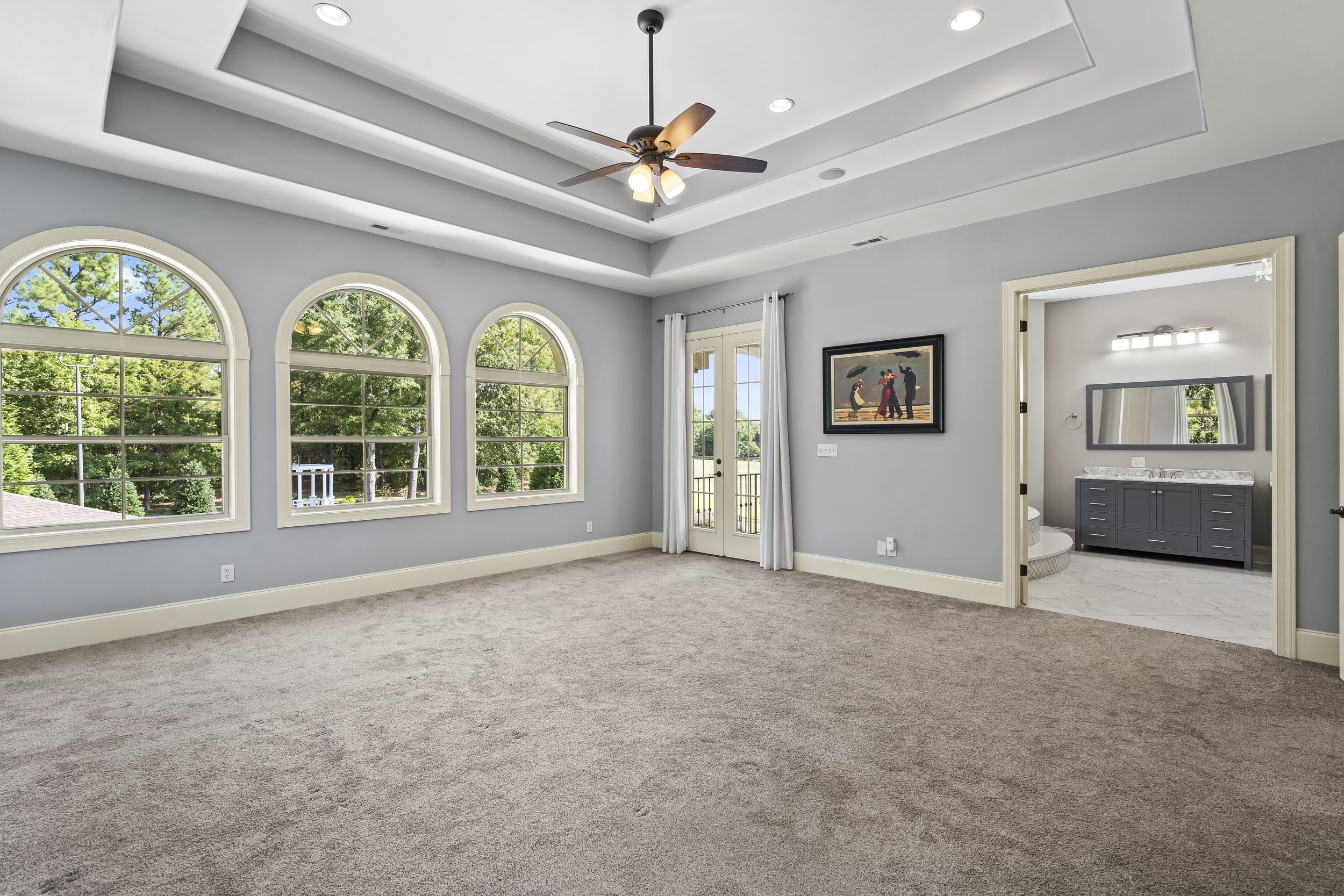 A spacious, bright bedroom with three large arched windows, a door leading to a balcony, a ceiling fan, and a view into an en-suite bathroom with a vanity, mirror, and light fixture.