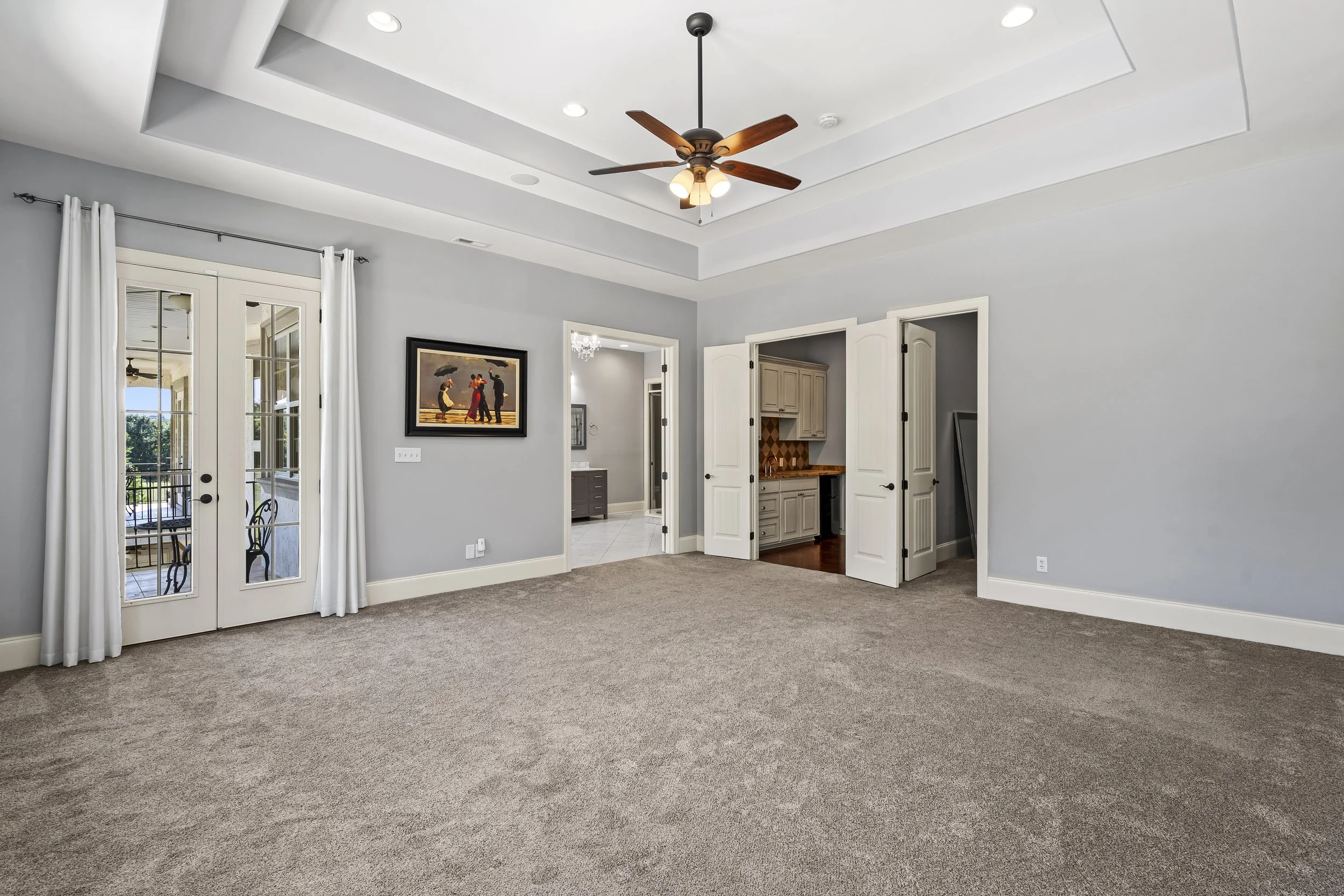 Empty living room with beige carpet, gray walls, double glass door leading to balcony, and open doorway to kitchen and hallway, ceiling fan with wooden blades, recessed lighting, and a wall-mounted artwork.