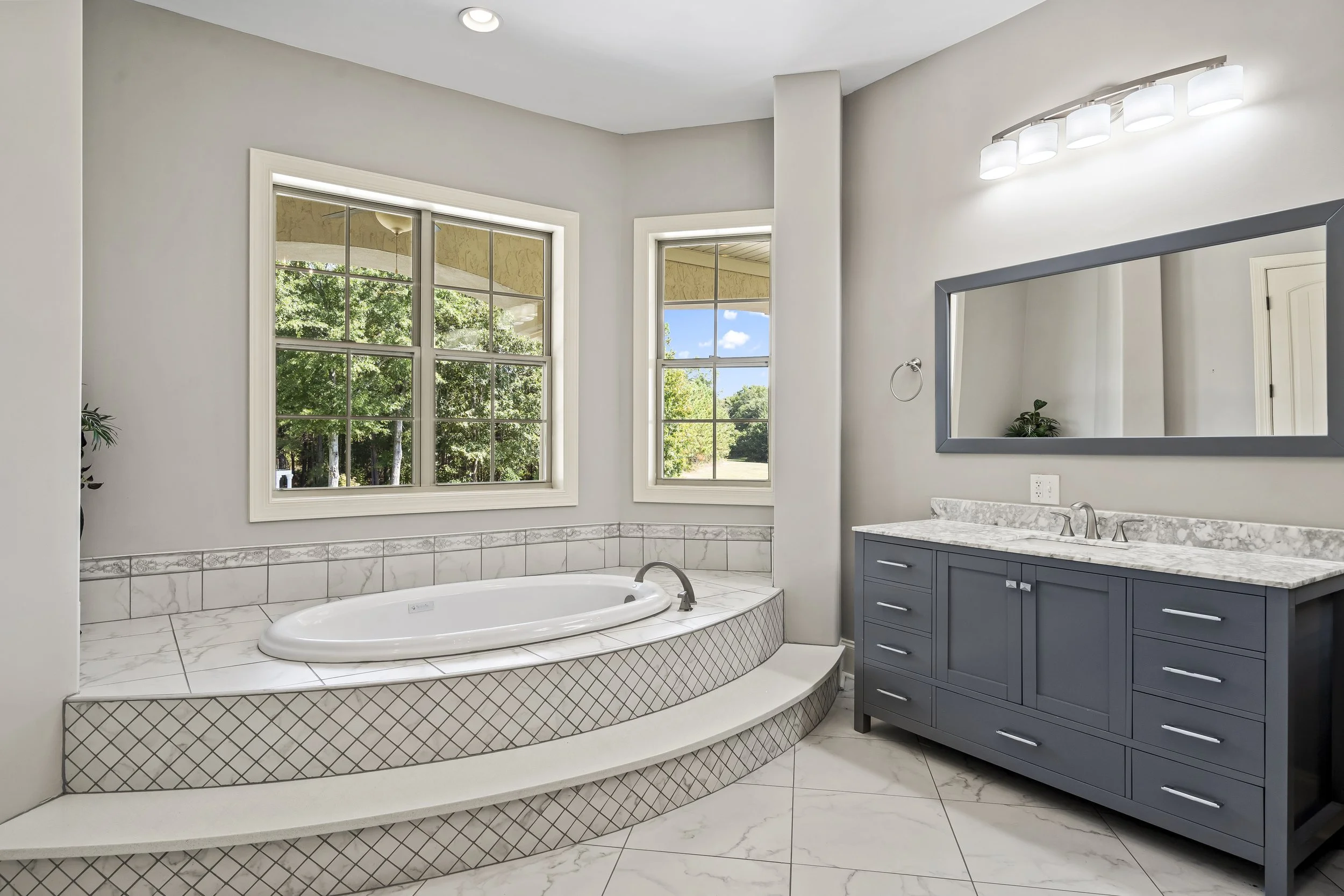 Bathroom with a bathtub under two large windows, a blue vanity with a marble countertop, a rectangular mirror, and a wall-mounted light fixture.