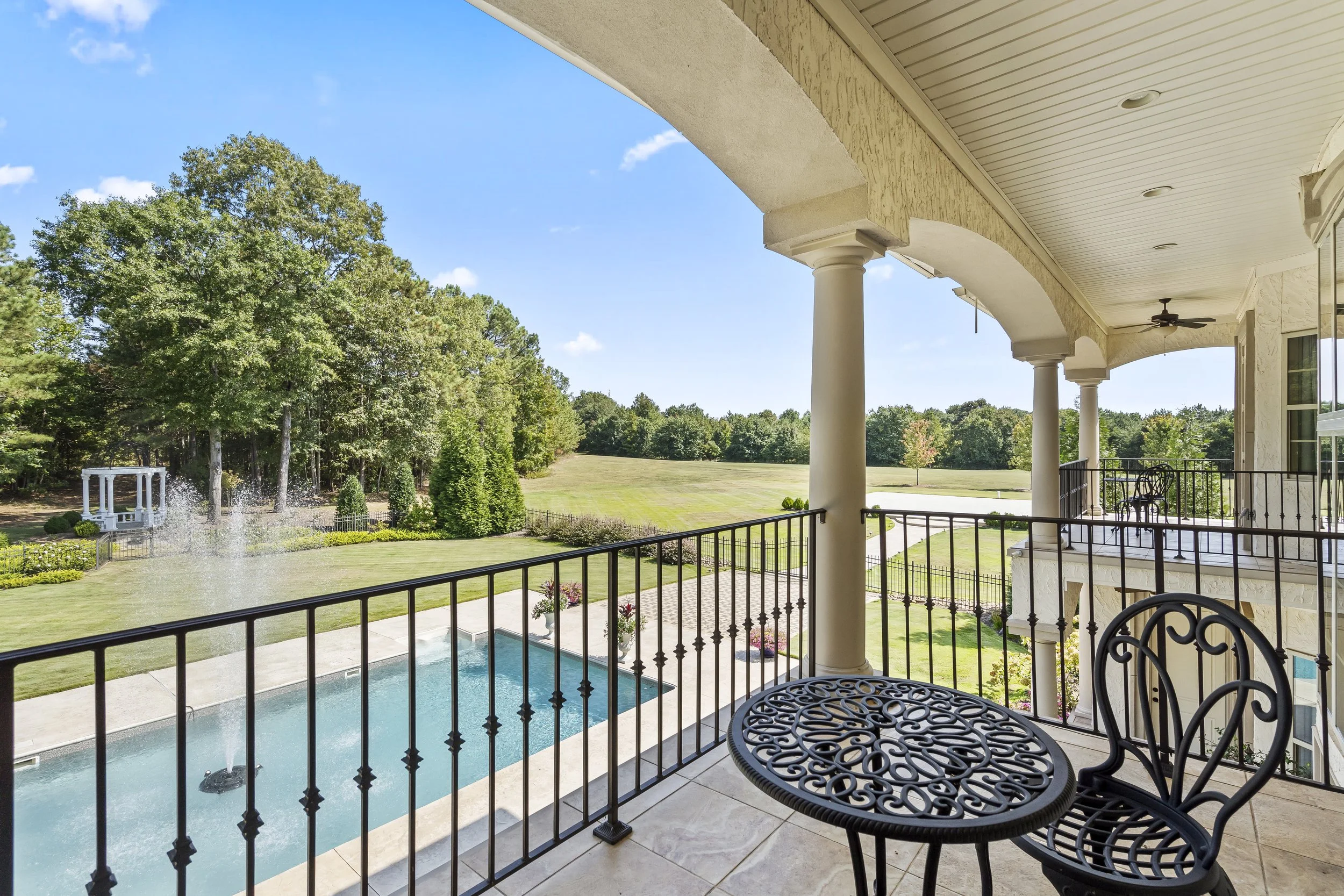 View from a balcony overlooking a backyard with a swimming pool, lush green trees, and a fountain on a clear, sunny day.