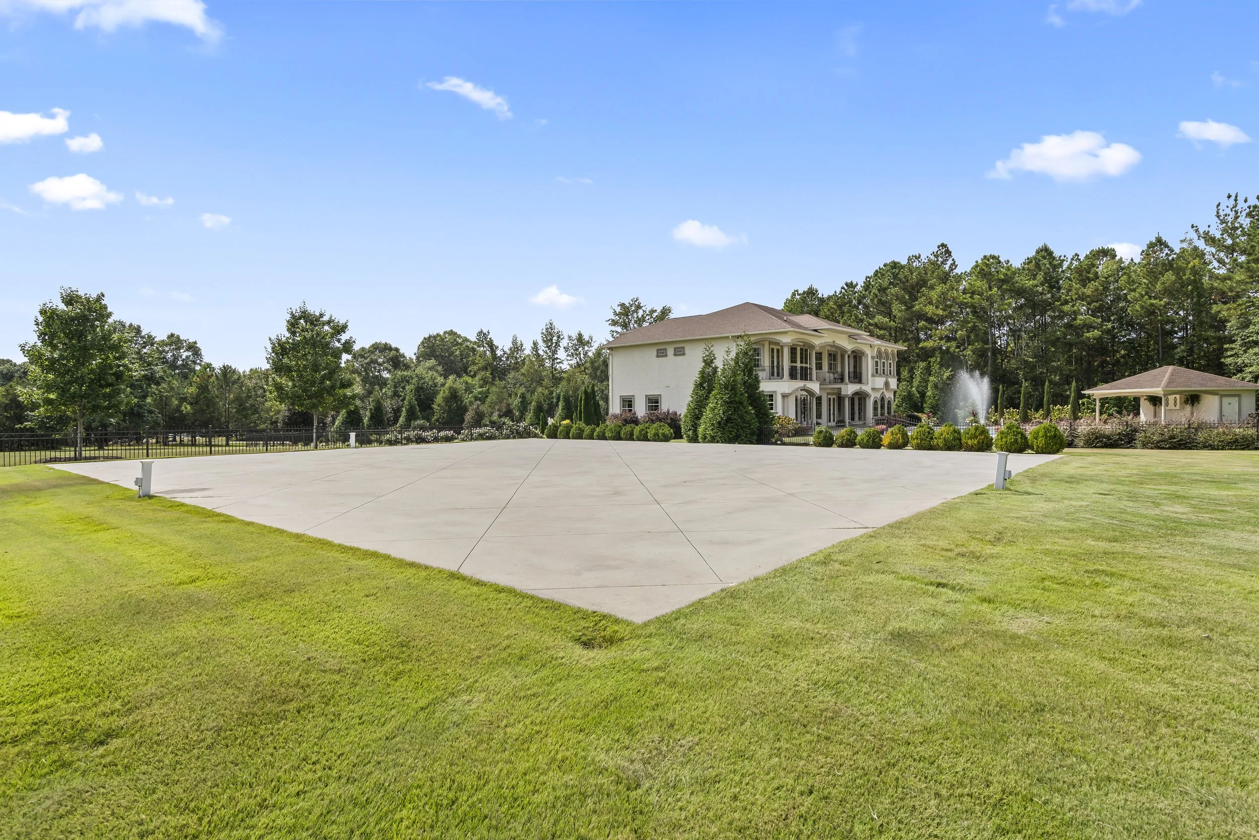 Large, empty concrete driveway leading up to a luxurious house with multiple balconies, surrounded by well-manicured lawn and trees, on a sunny day with blue sky and scattered clouds.
