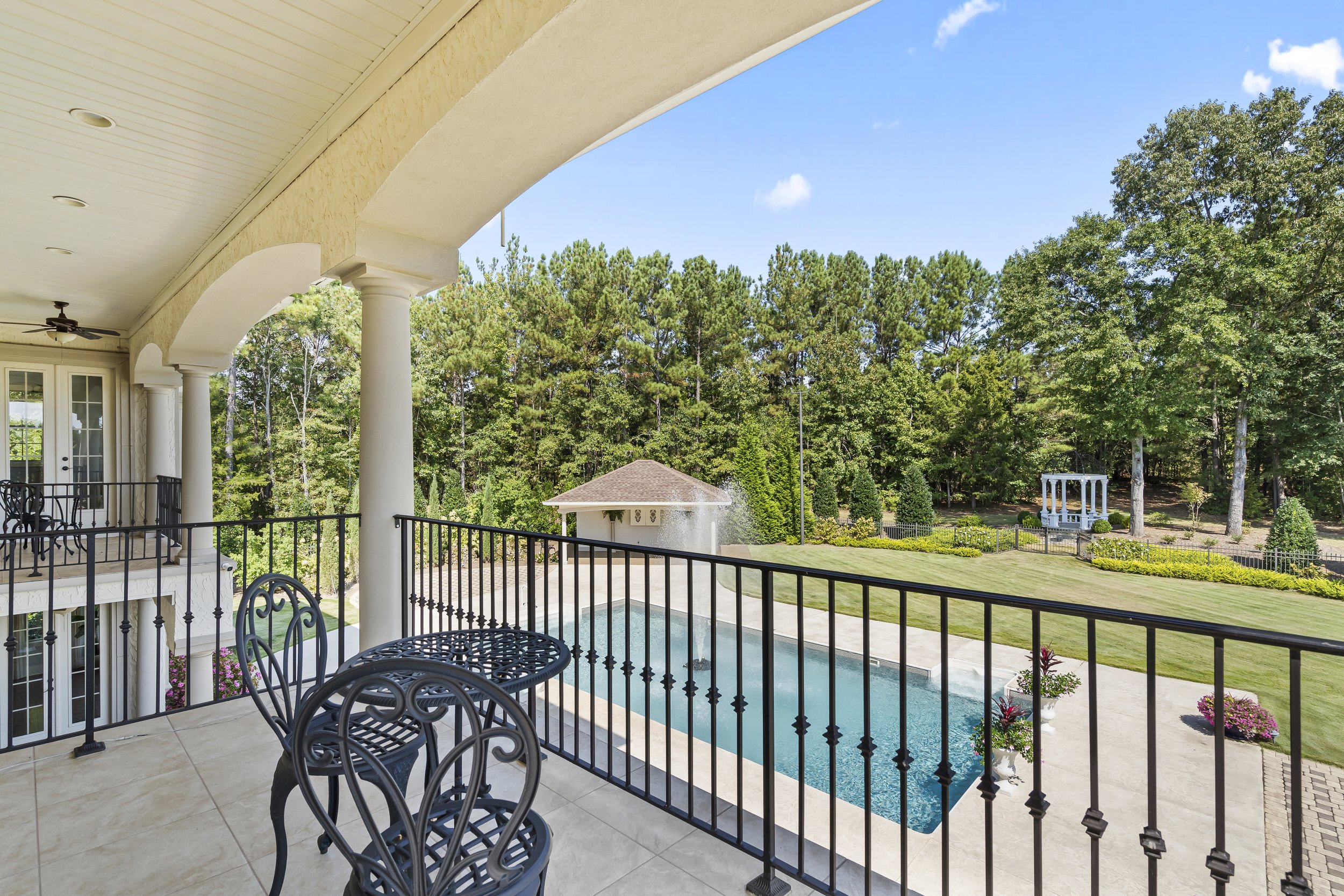 View of a backyard swimming pool from a covered balcony, with trees and a gazebo in the background.
