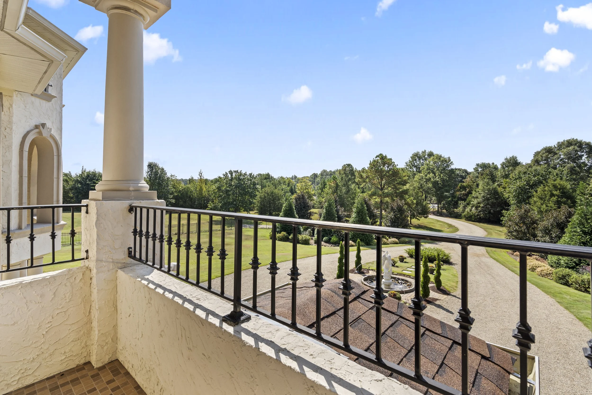 View from a balcony overlooking a garden with trees, a fountain, a statue, and a gravel driveway paved path under a blue sky with a few clouds.