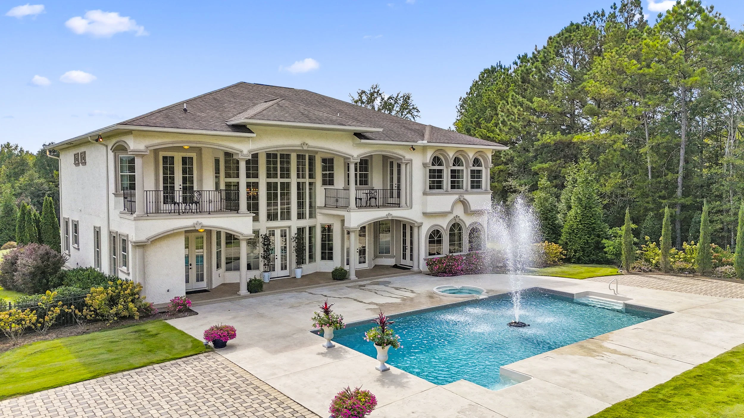 Large white two-story house with multiple windows, balconies, and a fountain in the backyard pool area. The yard has green grass, potted plants, and is surrounded by trees.