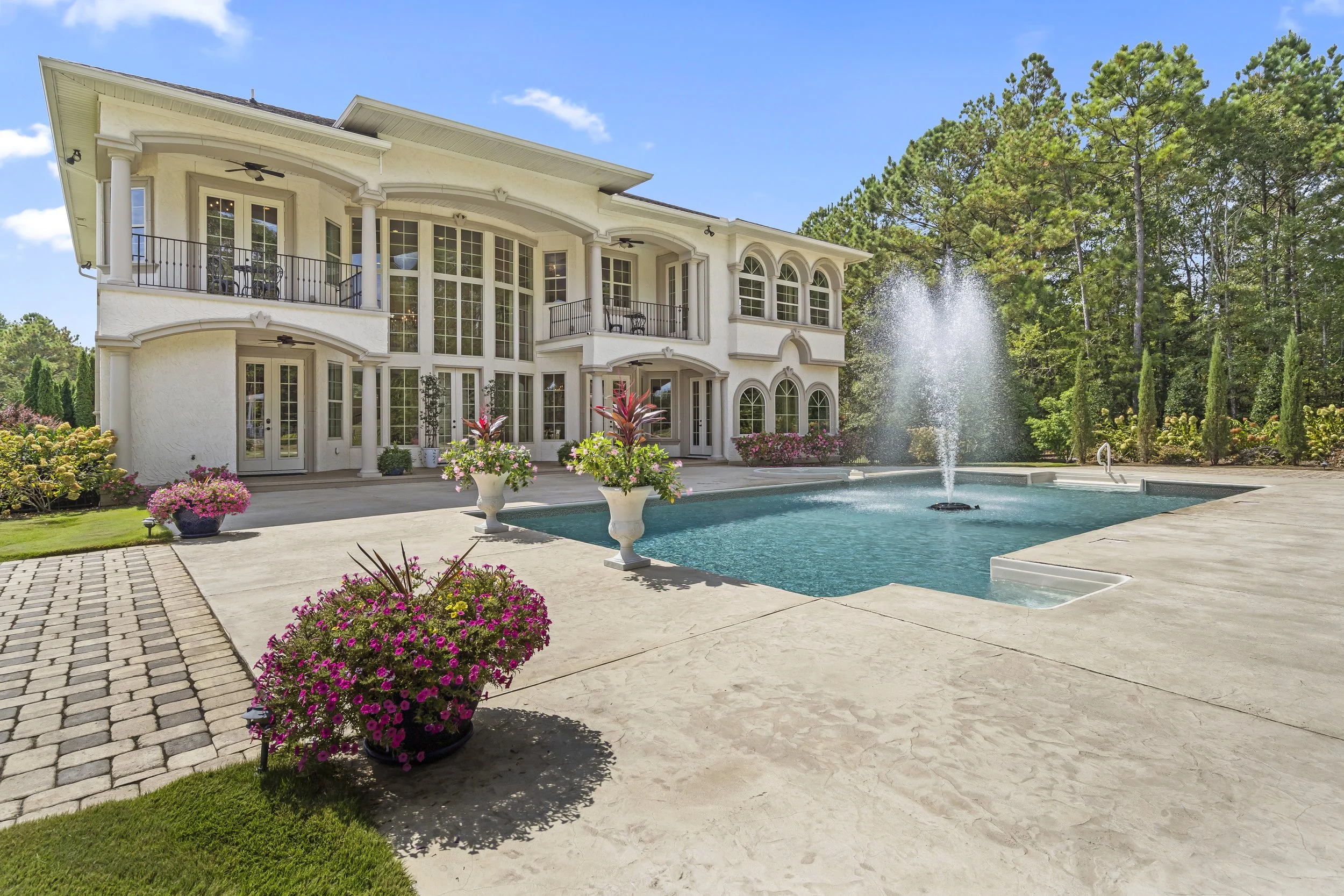 Large white house with rounded windows and balconies overlooking a backyard pool with a fountain, surrounded by colorful flowers and green trees.