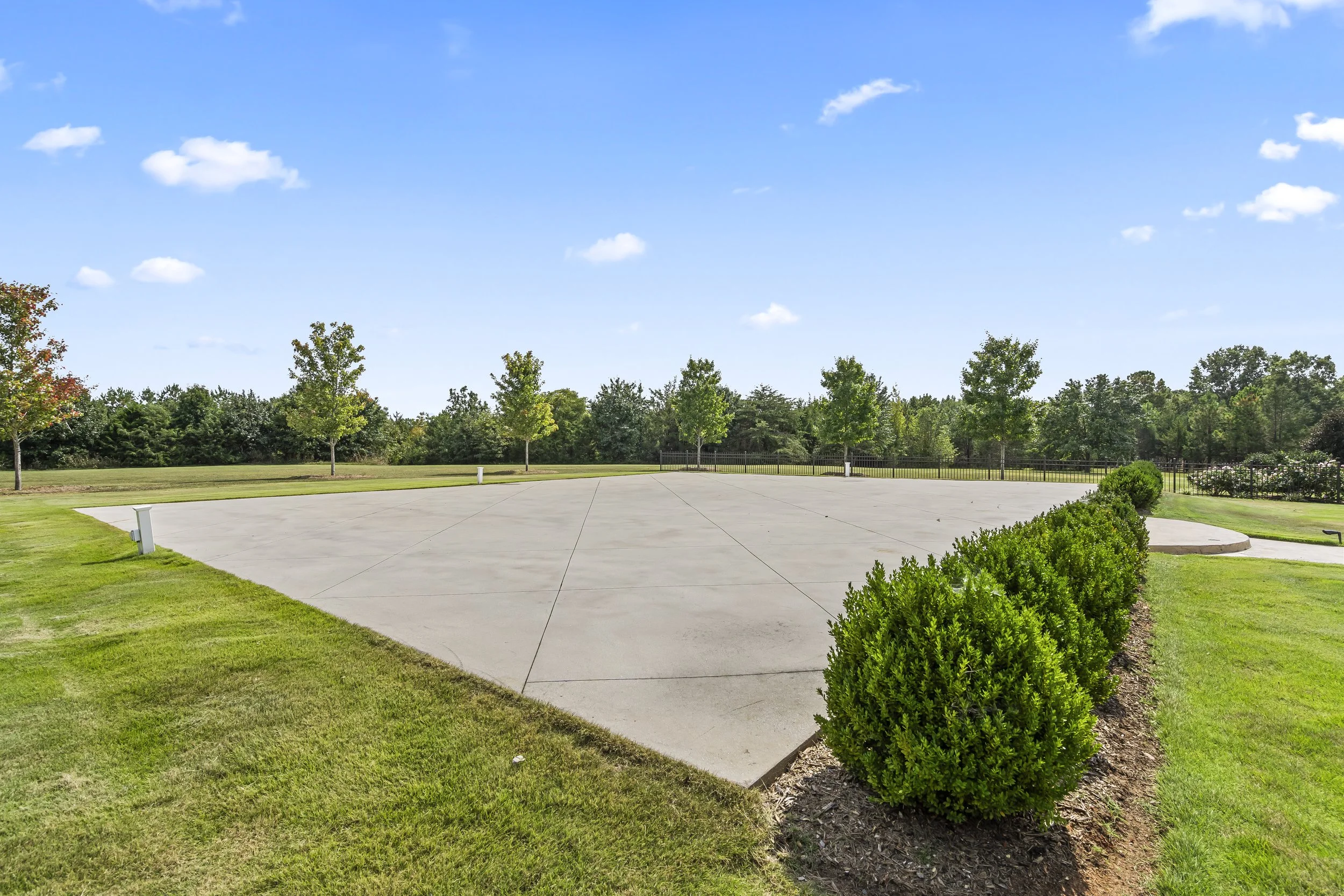 An empty concrete skate park or plaza with neatly trimmed bushes along the edge, surrounded by green grass and trees under a blue sky with scattered clouds.