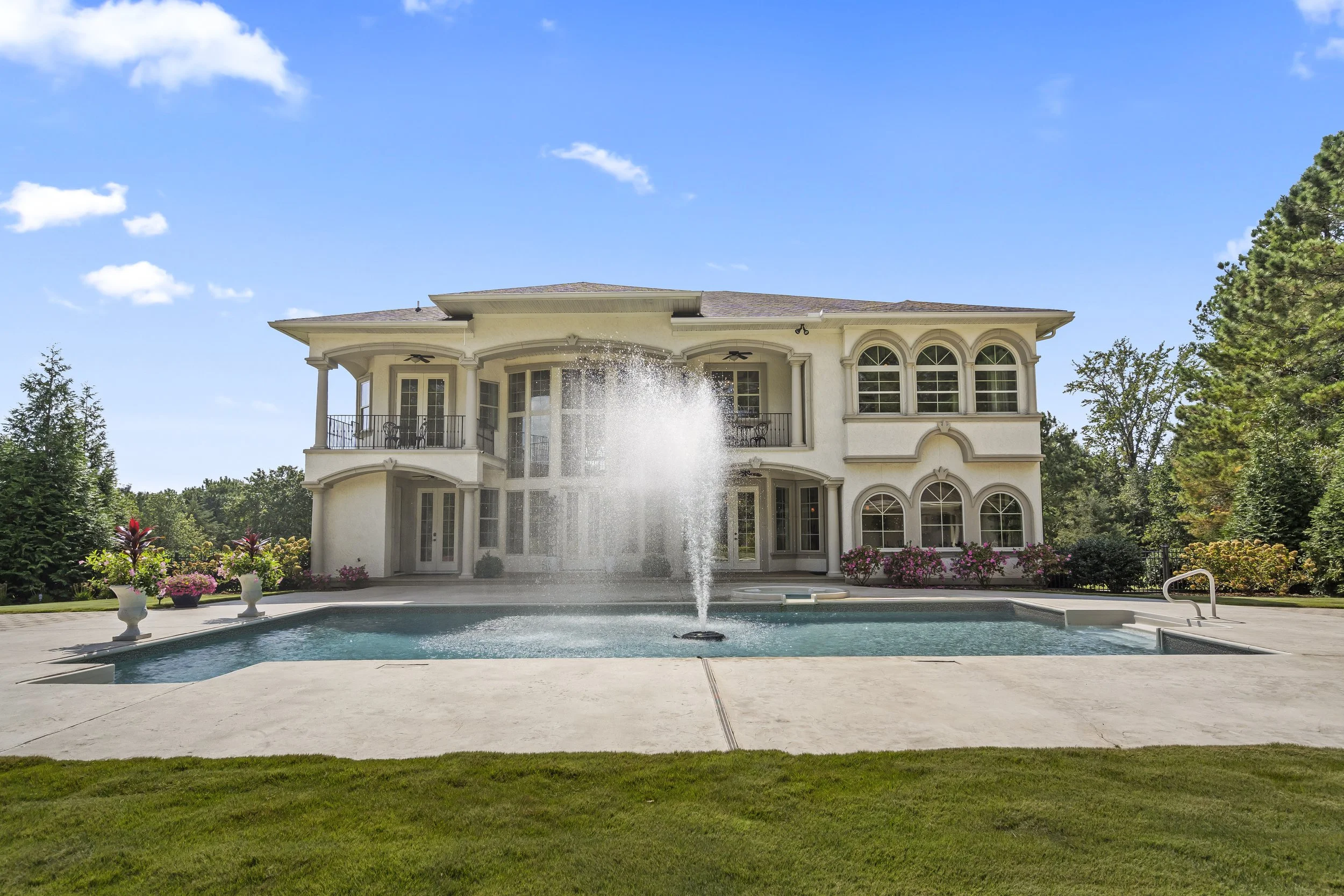 Large white mansion with multiple windows and balconies, a fountain in the swimming pool, and a well-maintained lawn and garden surrounding the property.