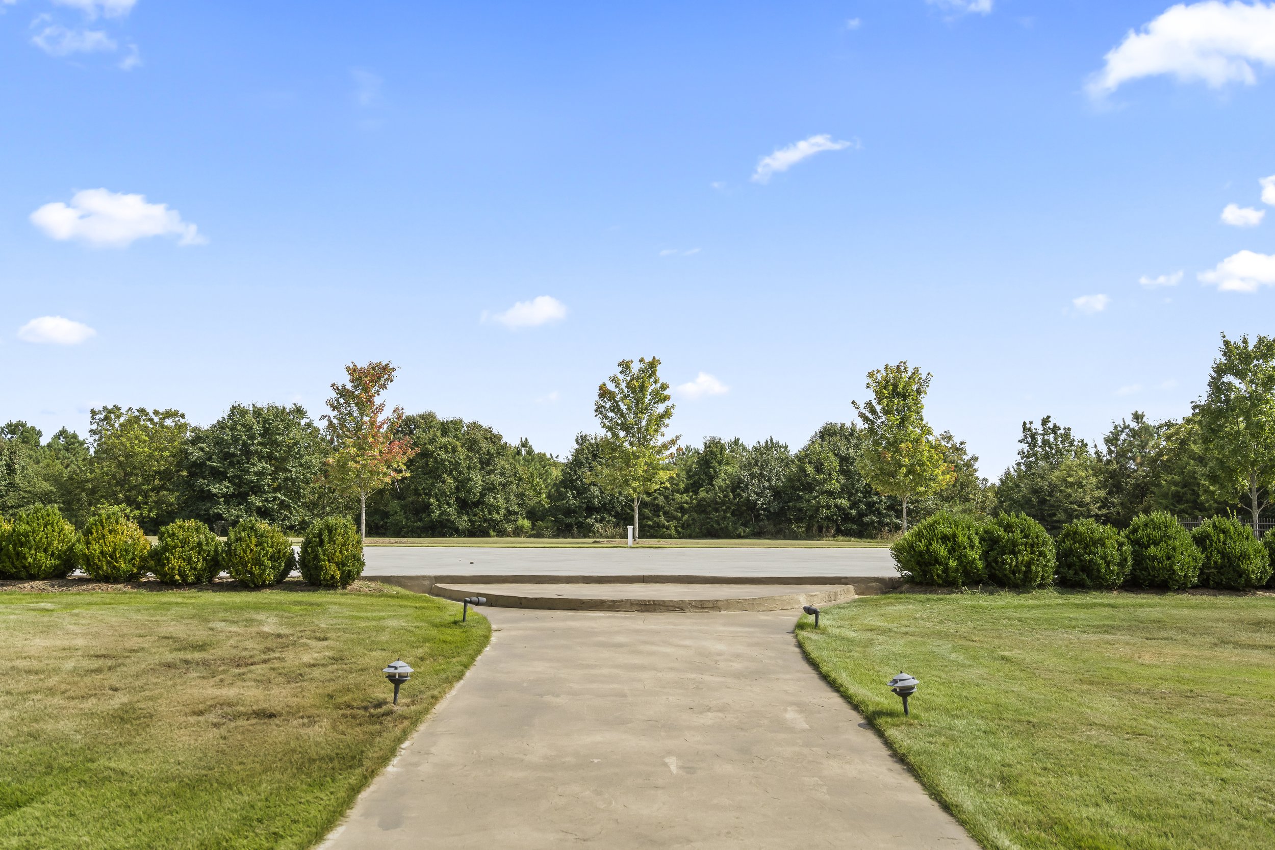 Concrete pathway leading to a small raised platform amidst well-maintained grassy lawns, with bushes and trees lining the background under a partly cloudy bright blue sky.