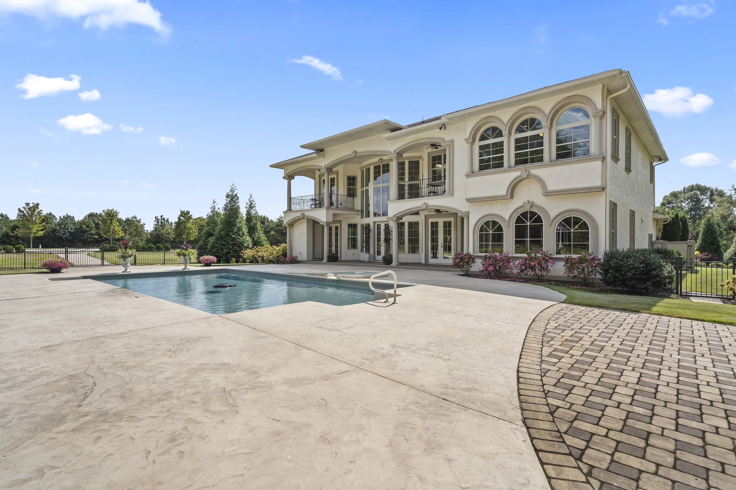 Large white house with multiple balconies and arched windows, surrounded by landscaped yard with pink flowers, a swimming pool, and a paved patio under a blue sky.