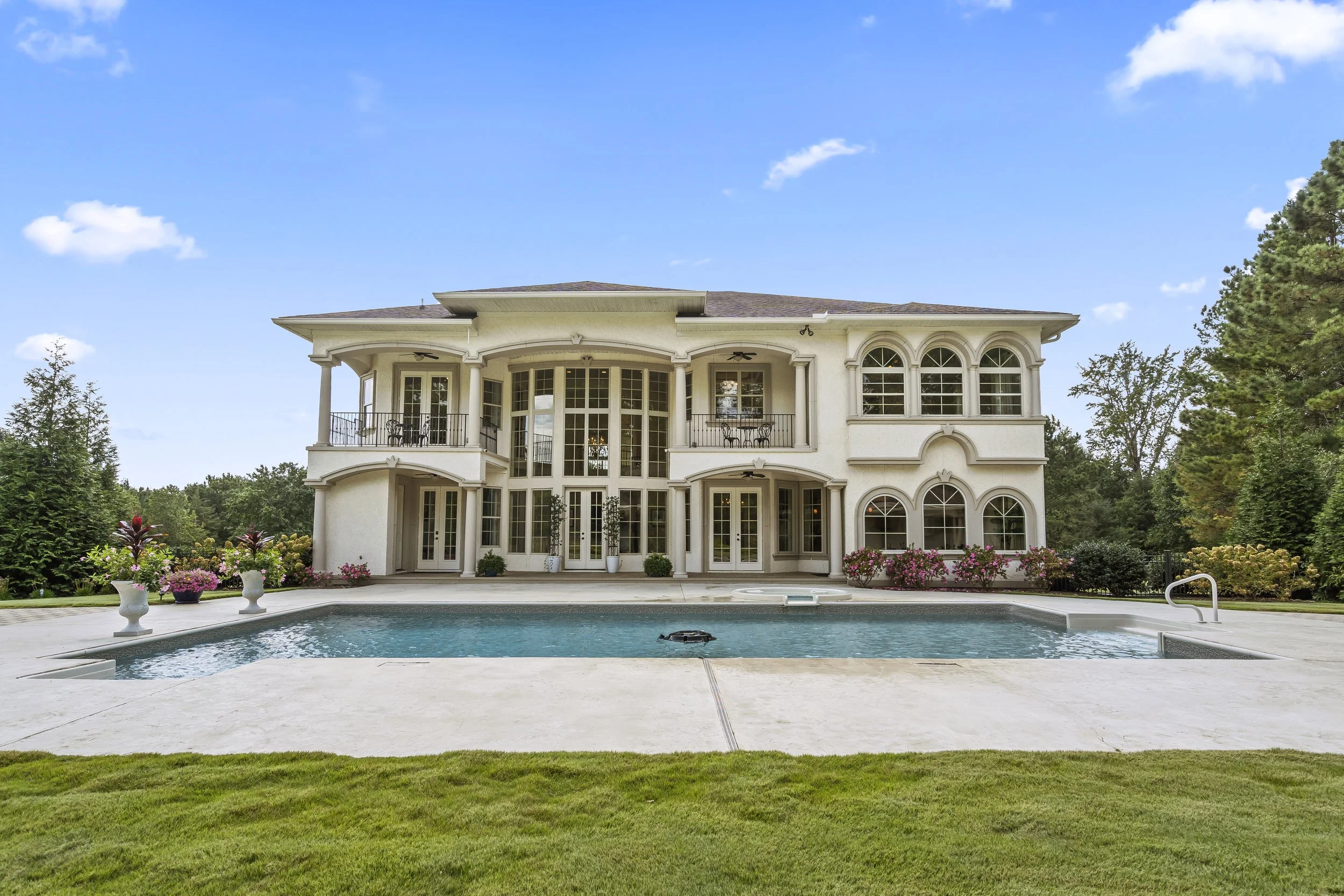 Large elegant white house with multiple balconies and arched windows, a backyard with a swimming pool, and landscaped garden with flowers and trees under a clear blue sky.