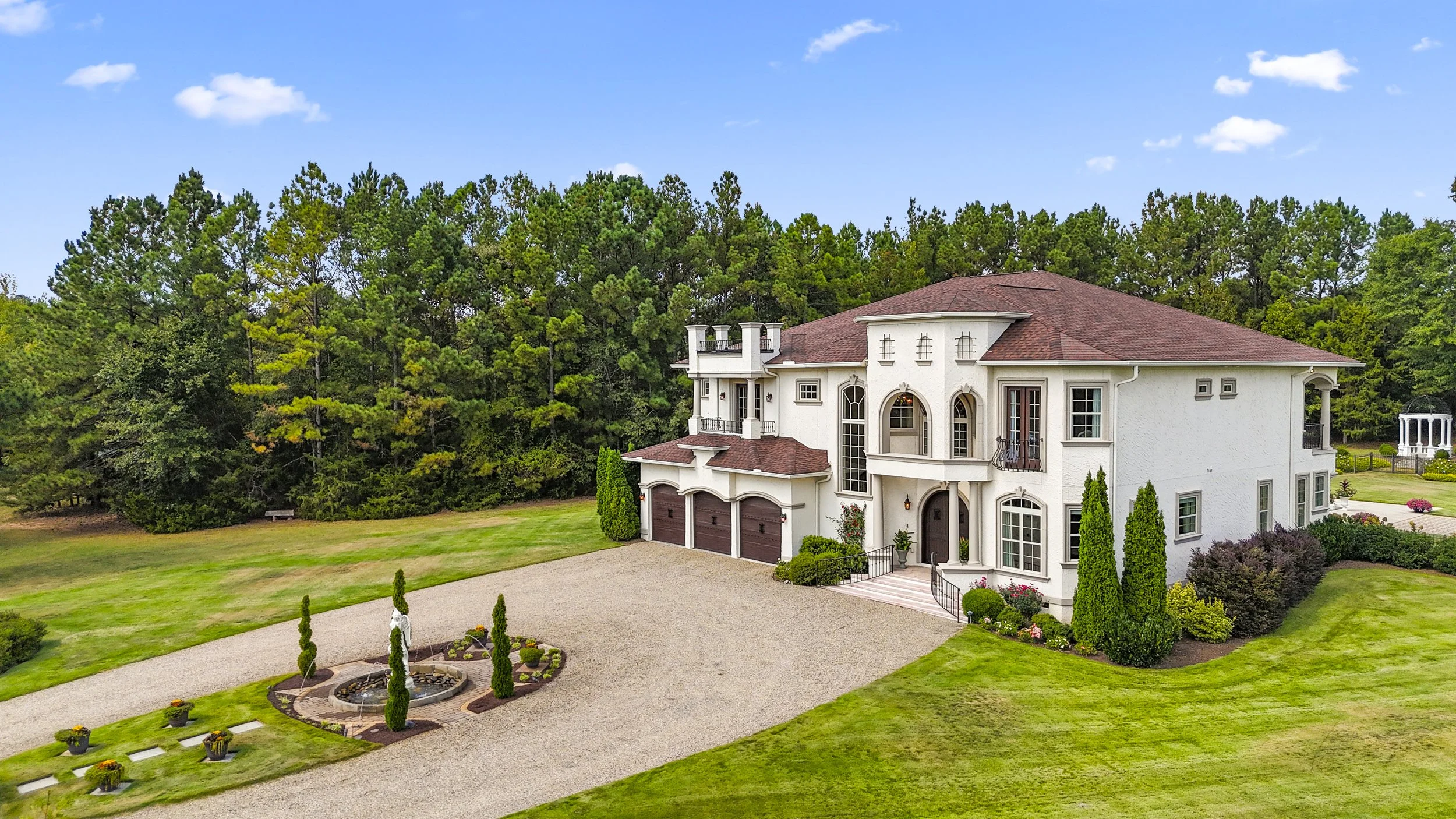 A large, luxurious white house with a red-tiled roof, surrounded by a well-maintained lawn and lush trees in the background. The house features arched windows, a balcony, and a three-car garage. There is a gravel driveway leading to the house with a 