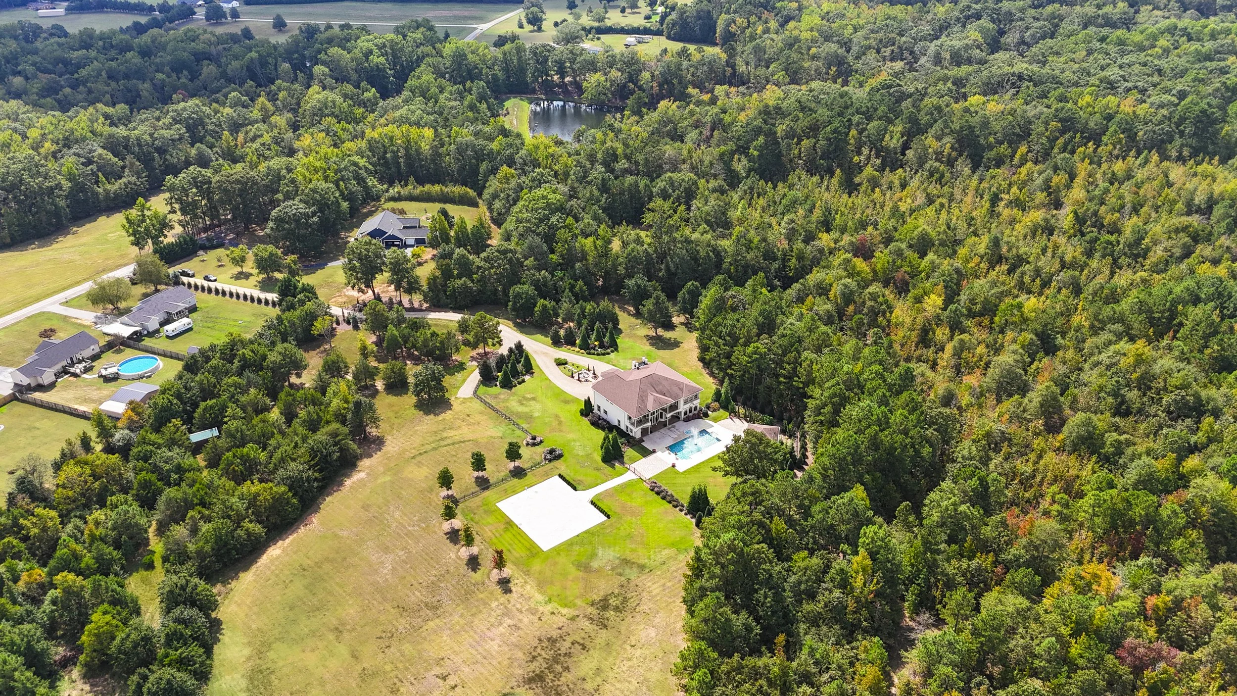 Aerial view of a residential area with houses, swimming pools, a tennis court, and surrounded by dense green trees and a forest, with a pond in the distance.