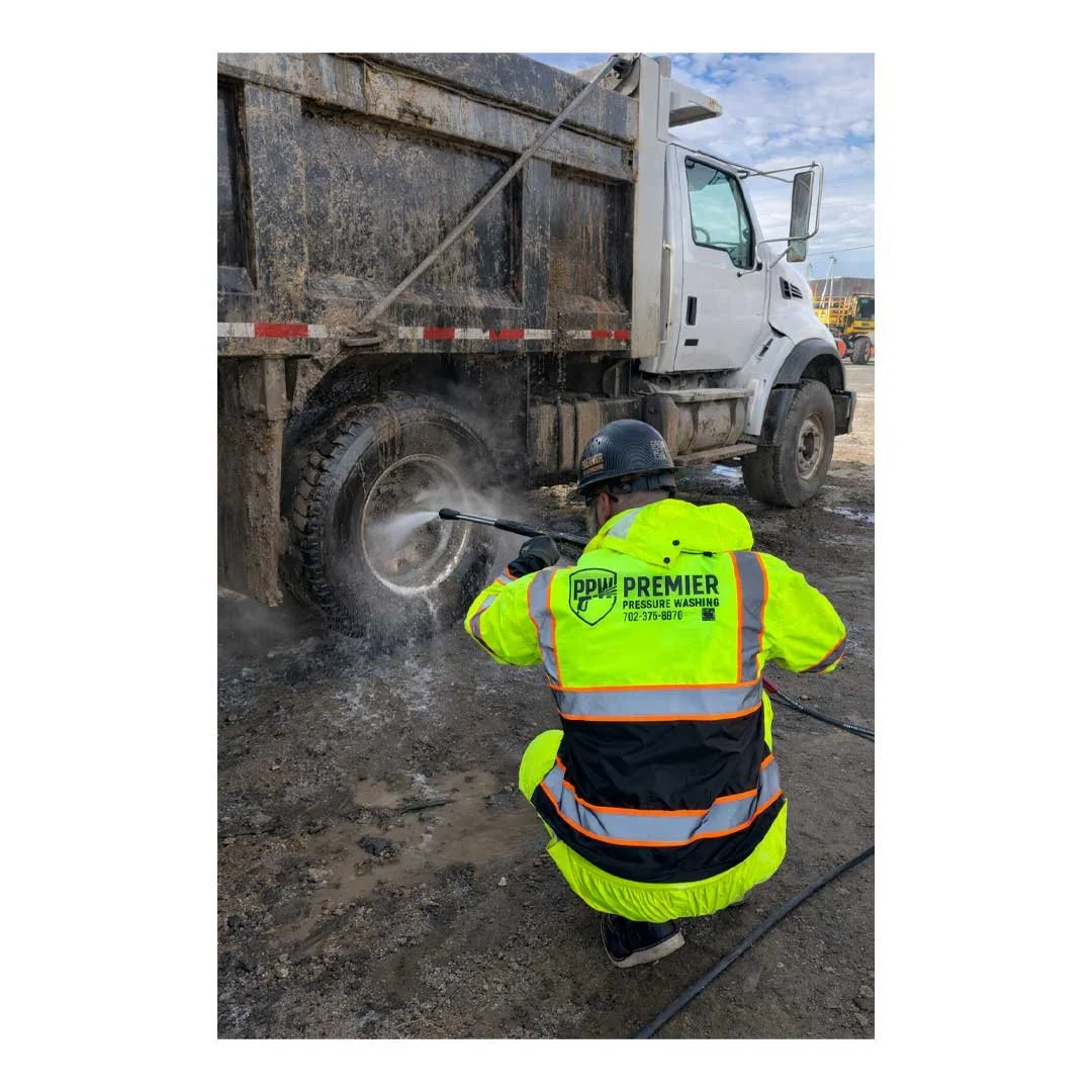 A worker in a high-visibility jacket and helmet is cleaning a dirty truck tire with a pressure washer at an outdoor job site.