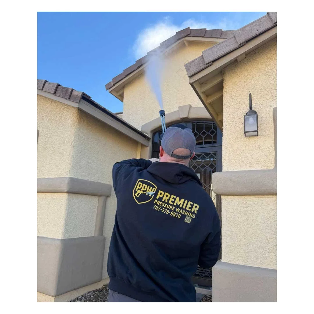A worker from Premier Pressure Washing is using a pressure washer to clean the exterior of a house. The worker wears a black hoodie with the company's logo on the back, and the house has beige stucco walls with a black front door.