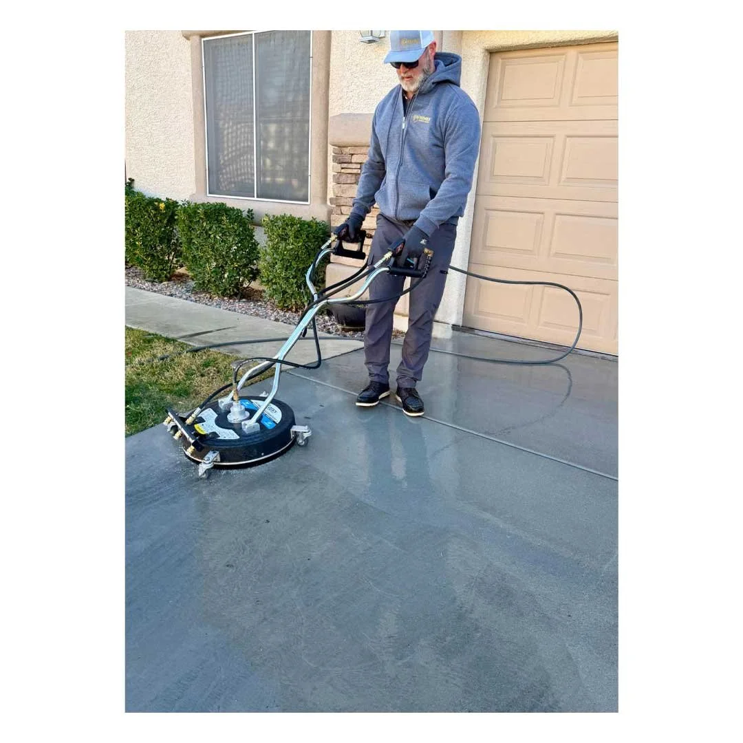 A man wearing a gray hoodie, gray pants, and a gray cap uses a concrete floor grinder outside a house with a tan garage door and a window with blinds, prepping the surface for sealing or coating.