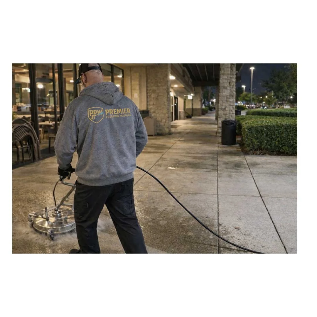 A worker cleaning an outdoor sidewalk with a power washer at night, wearing a gray hoodie with 'PPW Premier Pressure Washing' logo on the back.