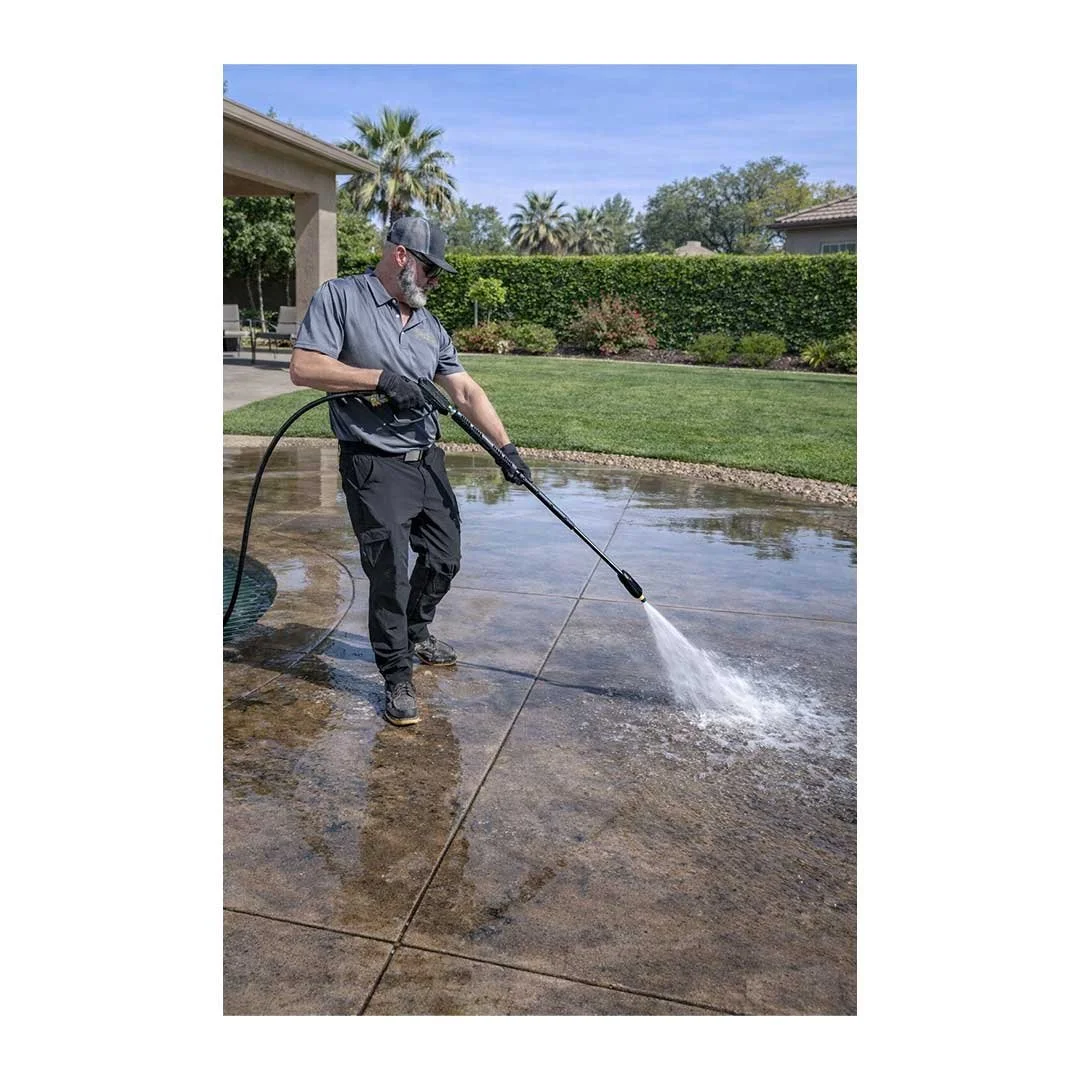 Man pressure washing concrete patio in backyard with green lawn and trees in the background.