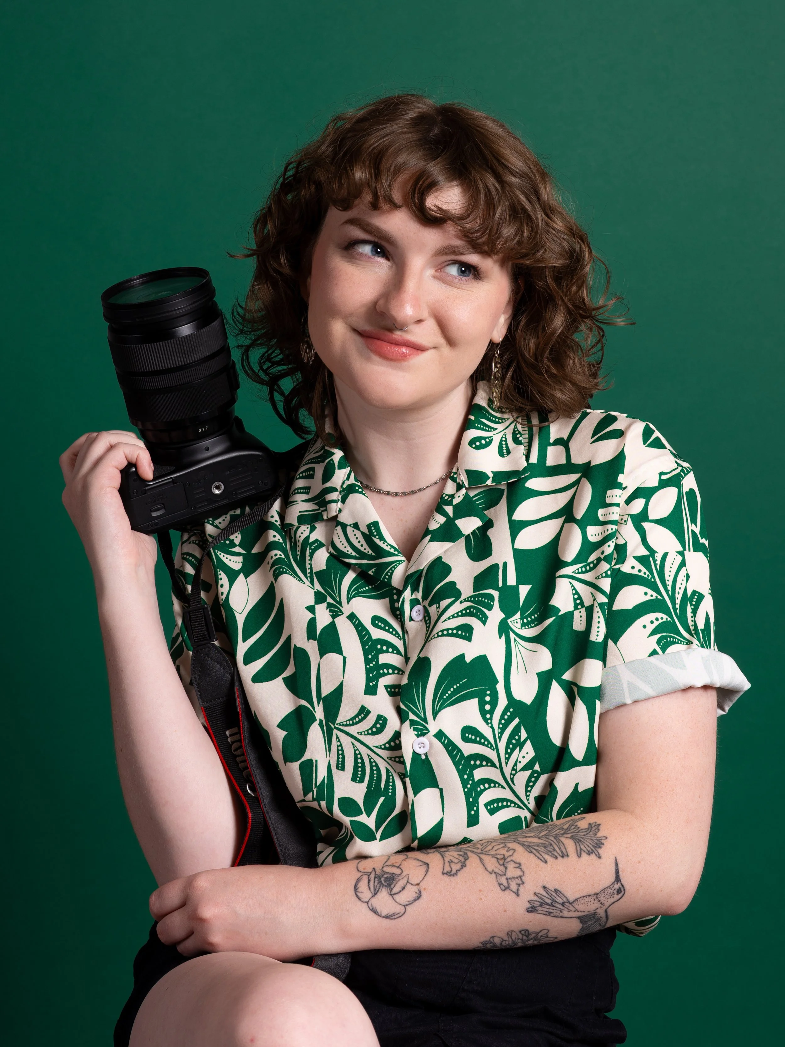 Hadley holding a camera to her shoulder, wearing a green and white patterned shirt, sitting against a green background.