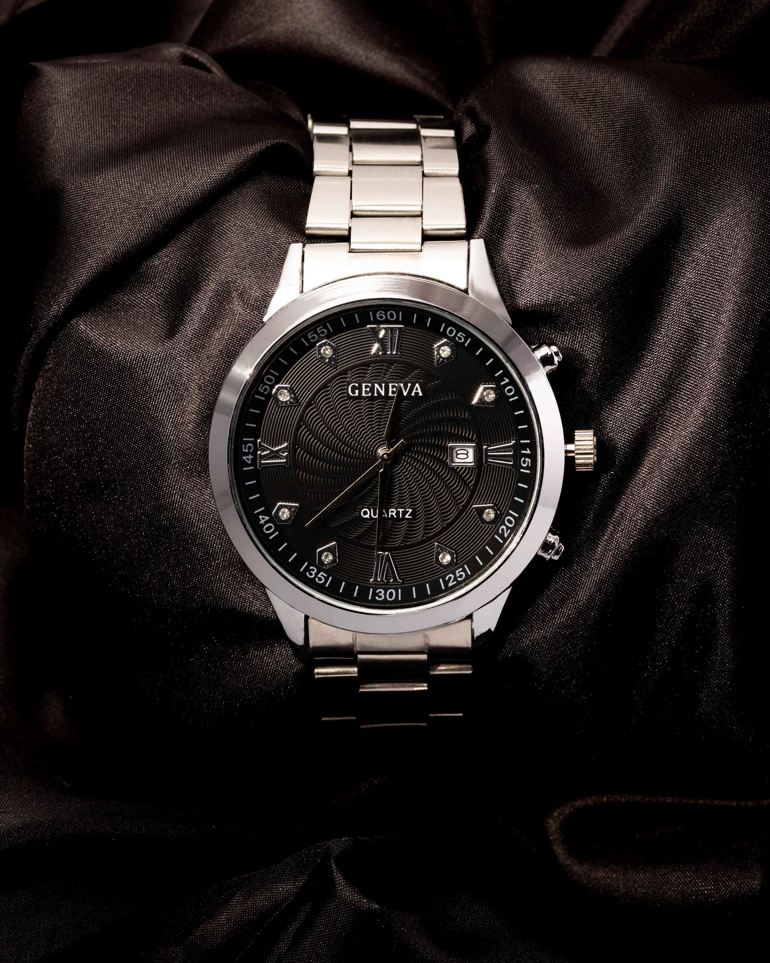Silver wristwatch with black dial, Roman numerals, diamond markers, date window, and metal band, resting on black satin fabric.