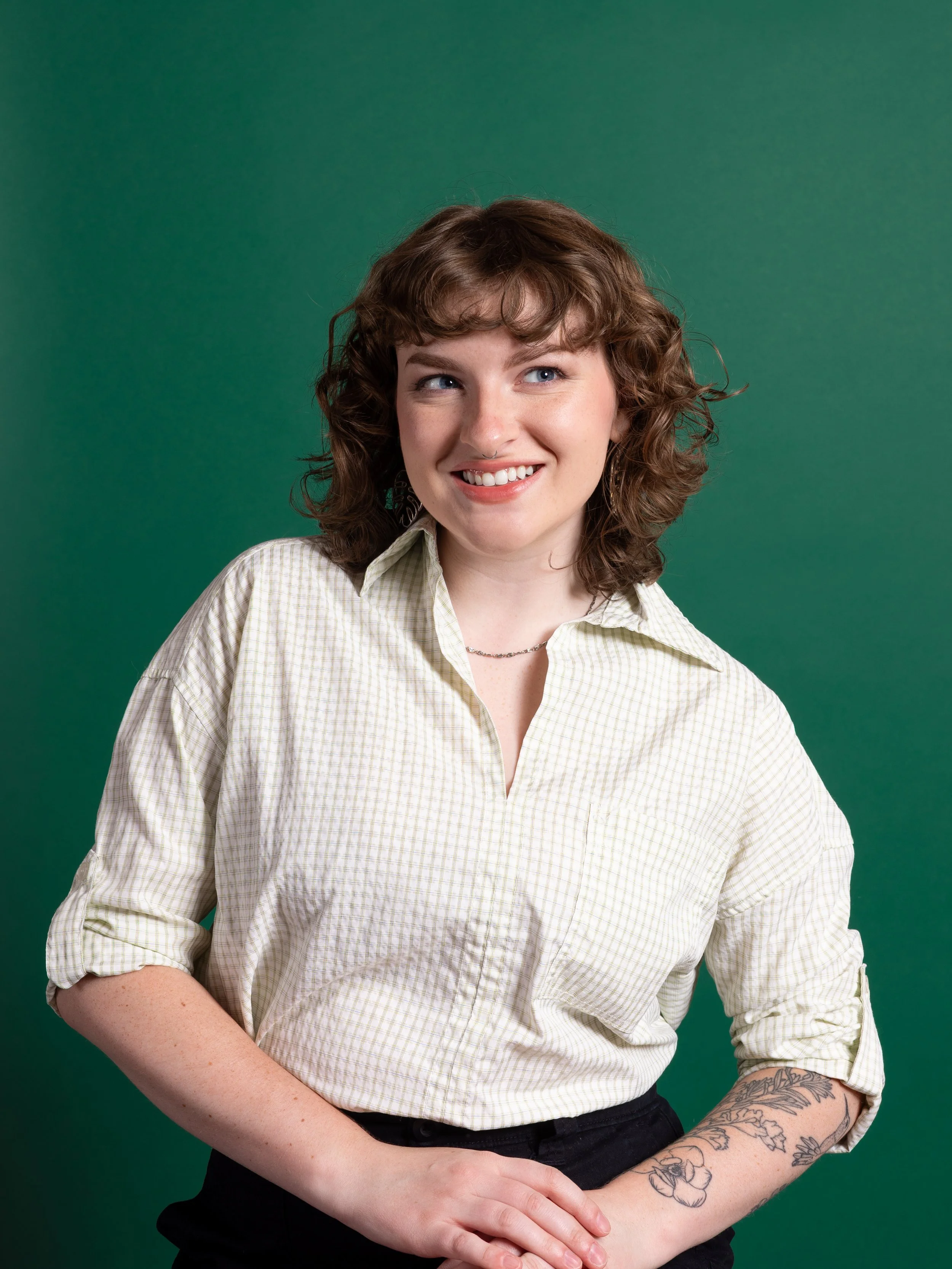 Hadley is smiling and looking to her right. She is wearing a light-colored checkered shirt with rolled-up sleeves, a necklace, and has tattoos on her left forearm. The background is solid green.