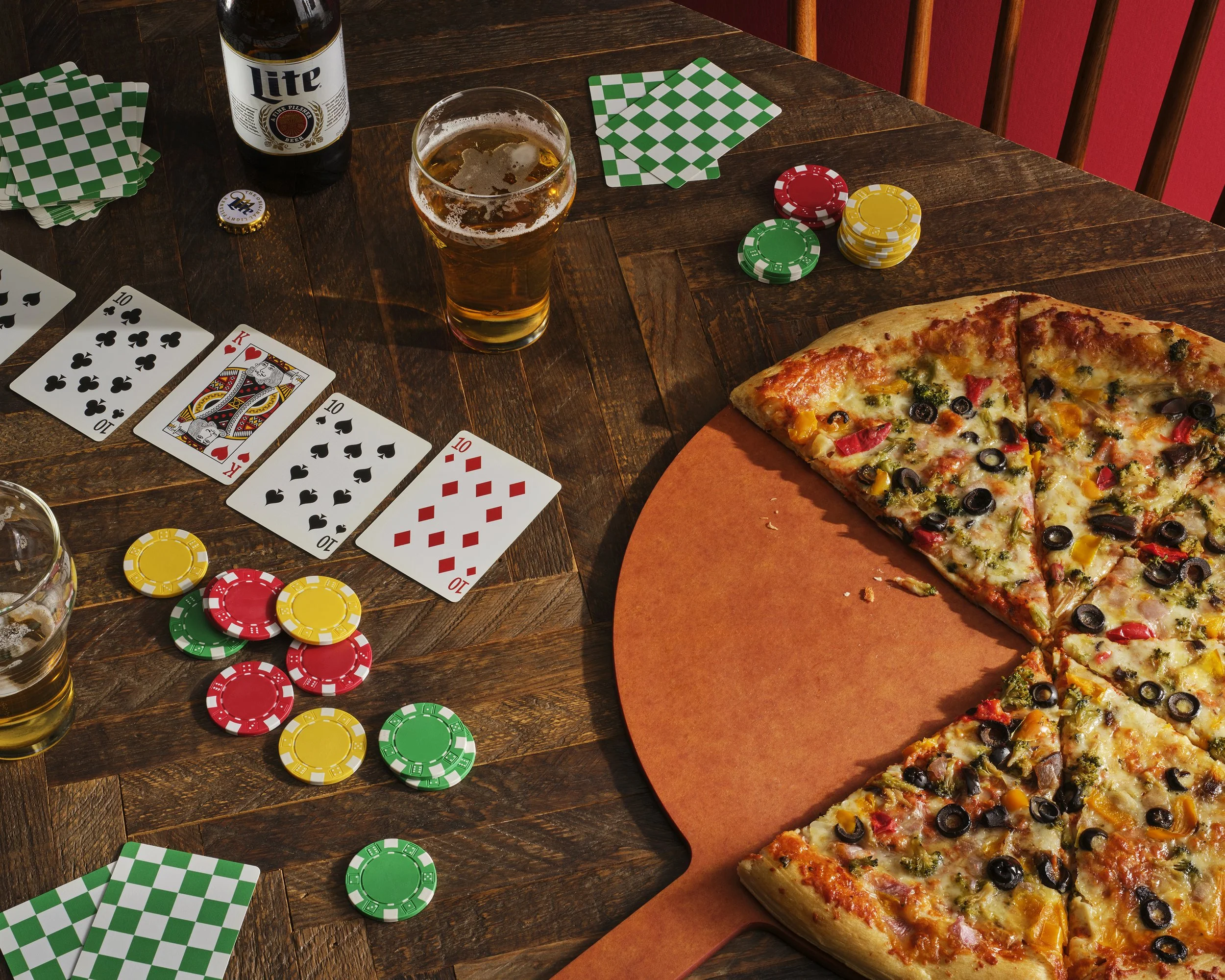 Food photography of a table set for a casual gathering with a pizza, playing cards, poker chips, and glasses of beer, in a cozy setting.