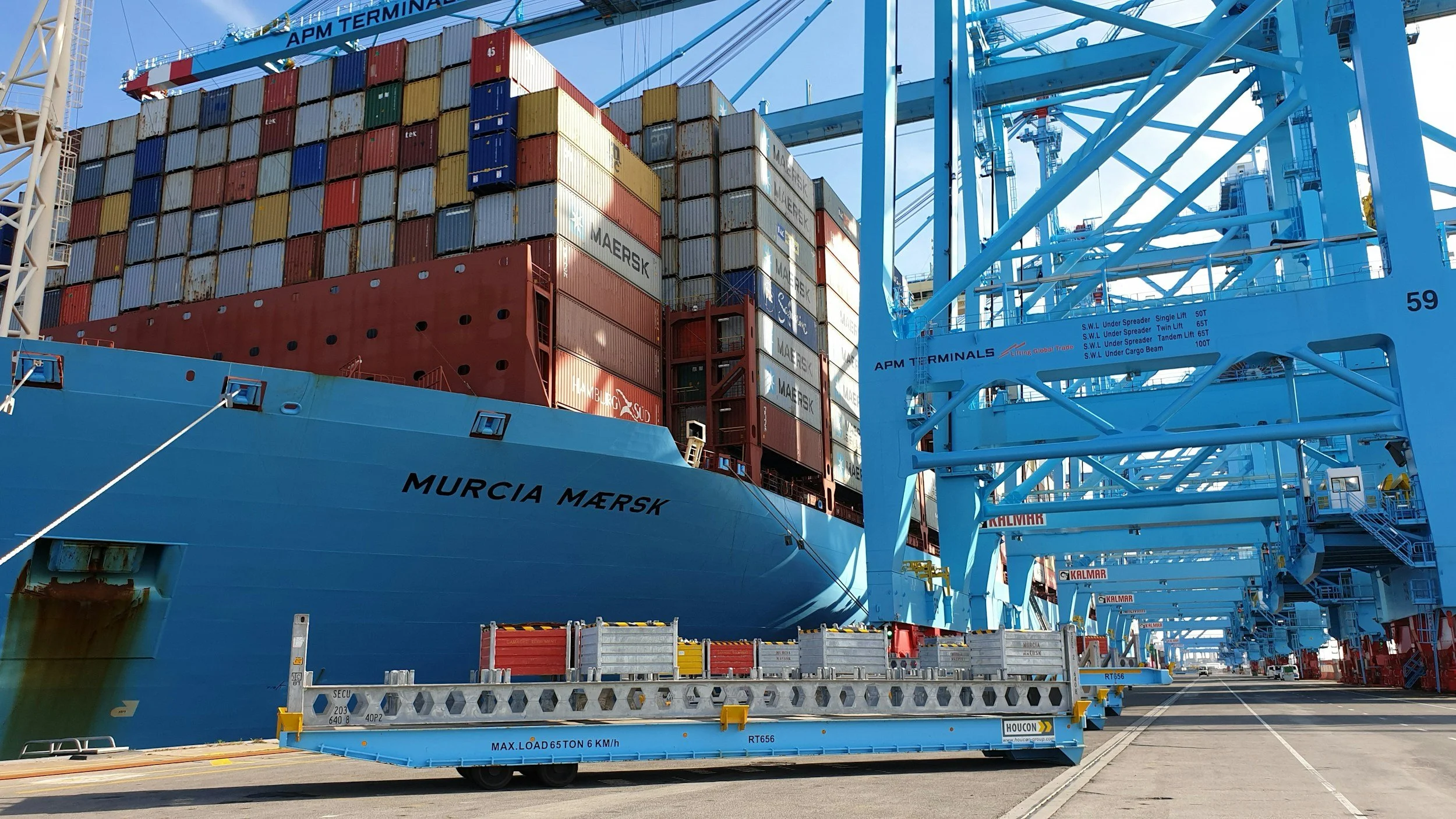 A large cargo ship named Murcia Mærsķ docked at a container terminal with numerous stacked shipping containers and large blue cranes for loading and unloading.