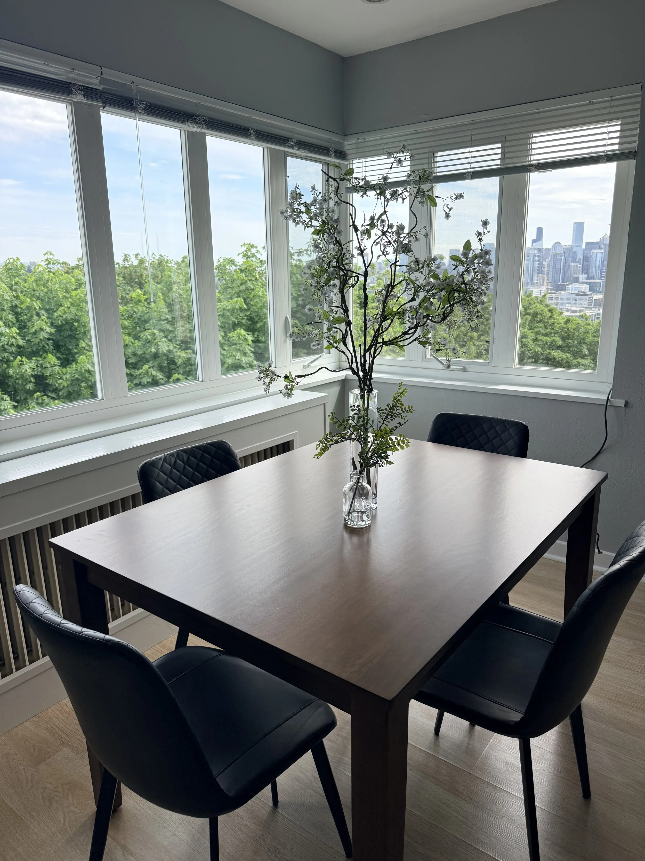 Modern dining room with a rectangular table, four black quilted chairs, a large window with a city view and a green canopy outside, and a decorative flower arrangement in tall glass vases.