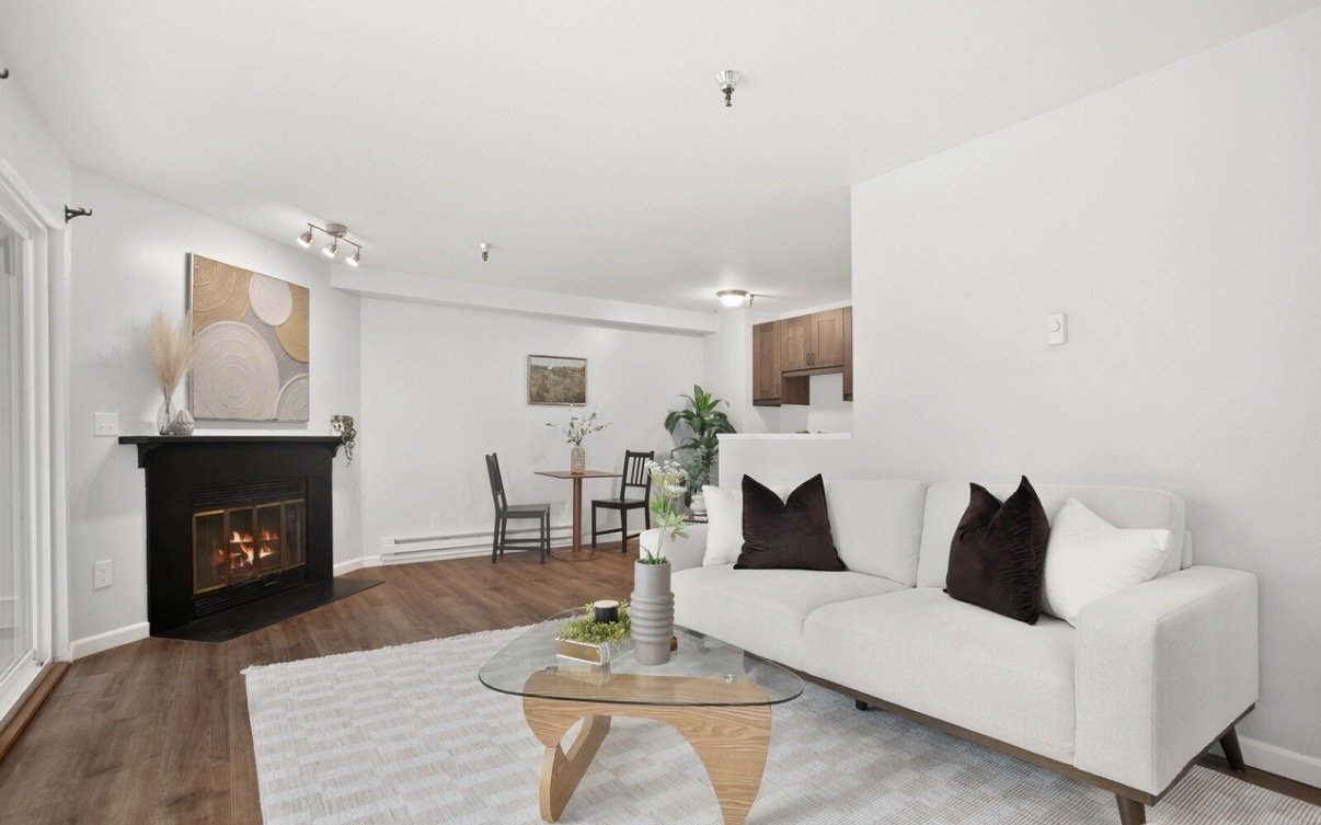 Living room with white walls, a white sofa with dark pillows, a glass coffee table, a fireplace with a black mantel, and a dining area with a round table and two chairs. There is a small plant near the sofa, artwork on the walls, and hardwood floors.
