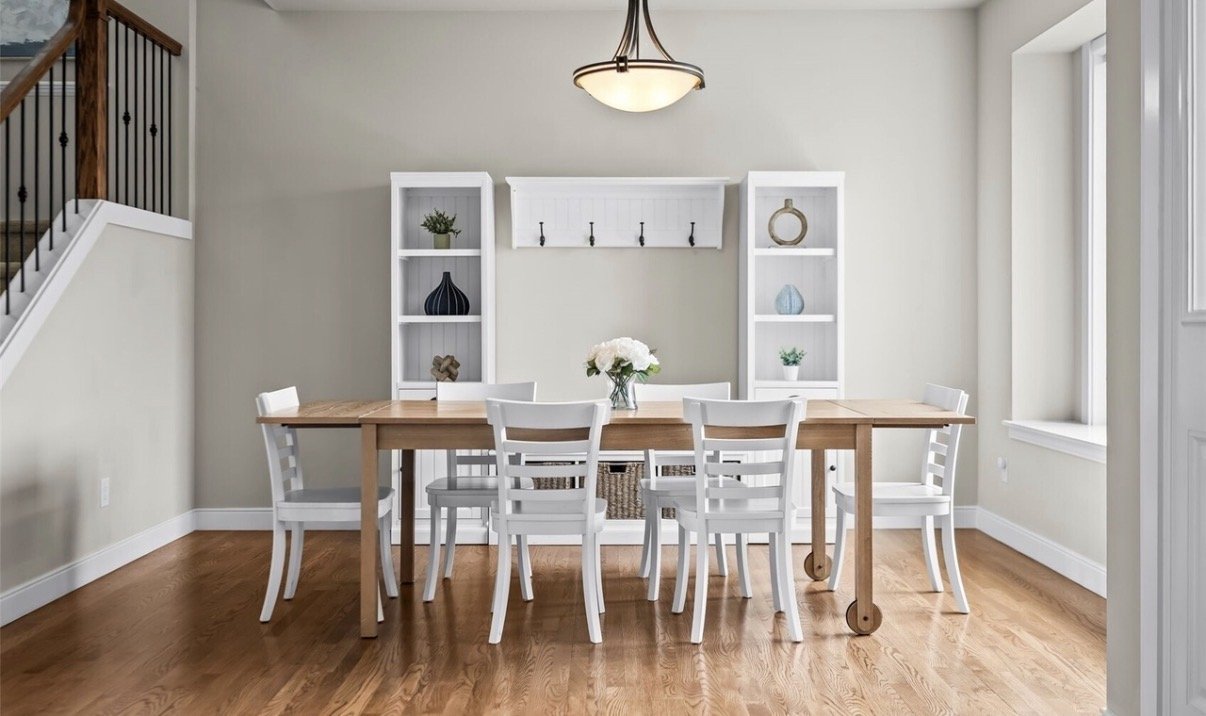 Dining room with wooden table and six white chairs, beige walls, two white bookshelves with decorative items, a flower arrangement in the center of the table, window on the right, ceiling light fixture, staircase on the left.