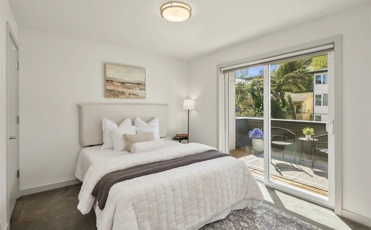 A bedroom with a white bed, multiple pillows, a blanket, a bedside lamp, a sliding glass door leading to a balcony with outdoor chairs and plants, and a minimalist art piece on the wall.