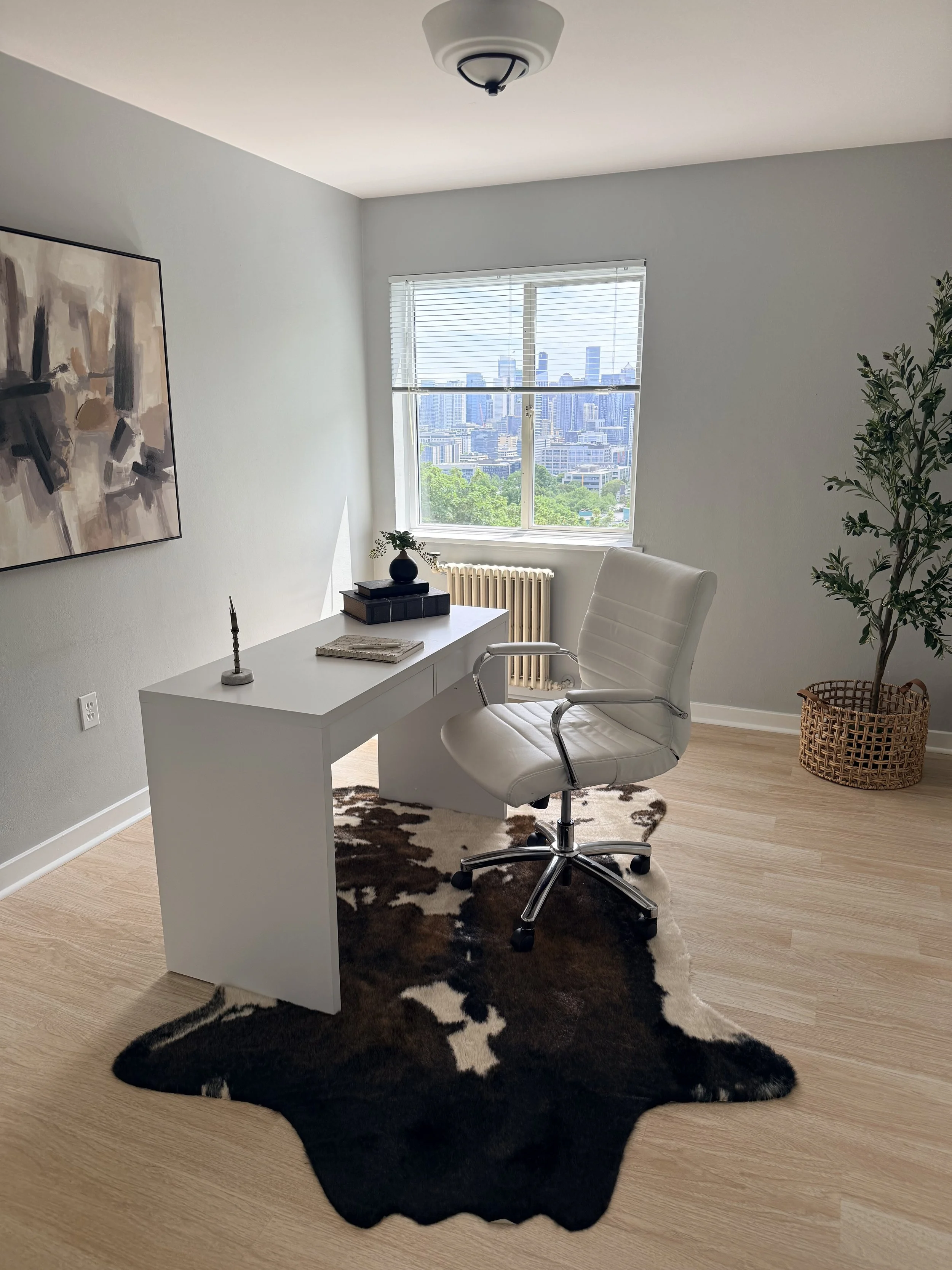 Modern office with white desk, white leather ergonomic chair, black and white cowhide rug, abstract wall art, small black vase with greenery, window with city skyline view, potted plant in basket.