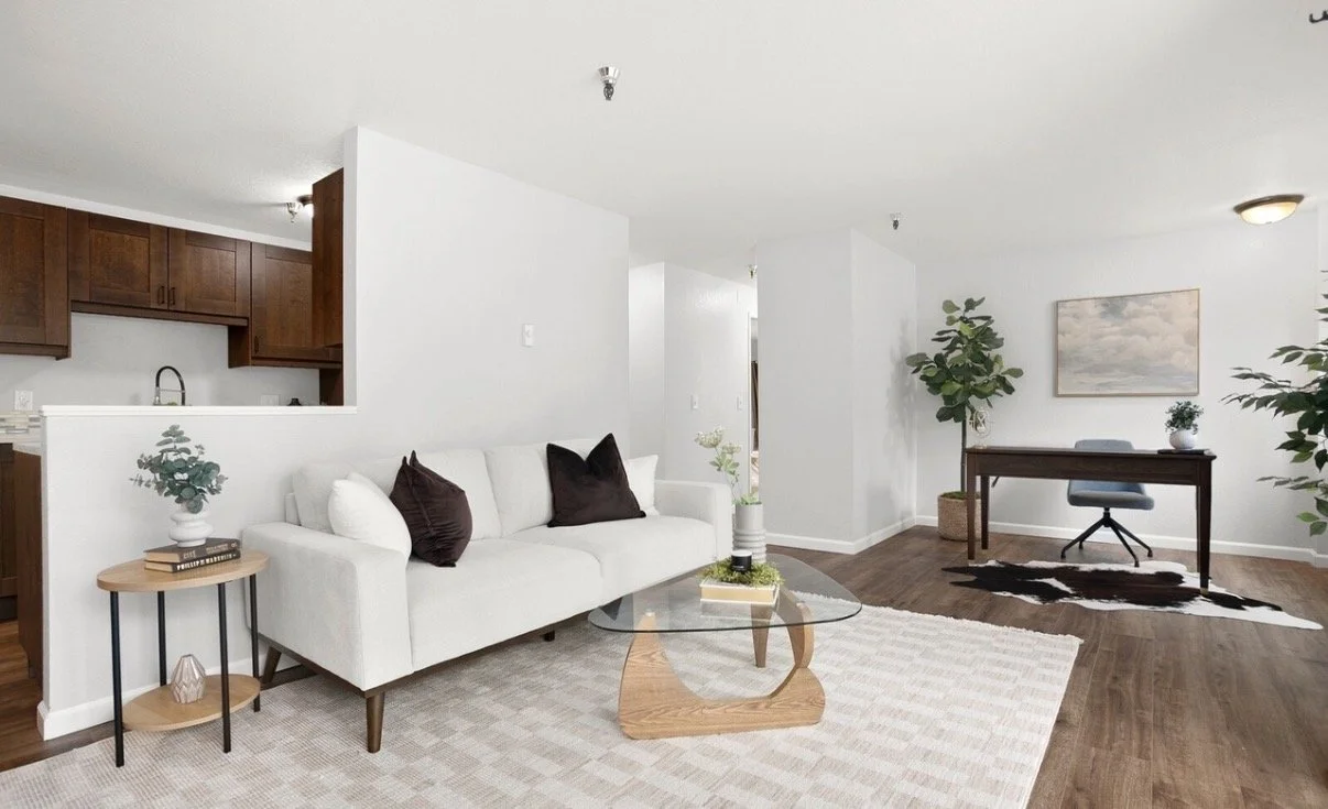 Living room with white walls and hardwood floors, white sofa with dark pillows, glass coffee table, small side table with plant, and a desk with a chair, framed artwork, and large indoor plants.