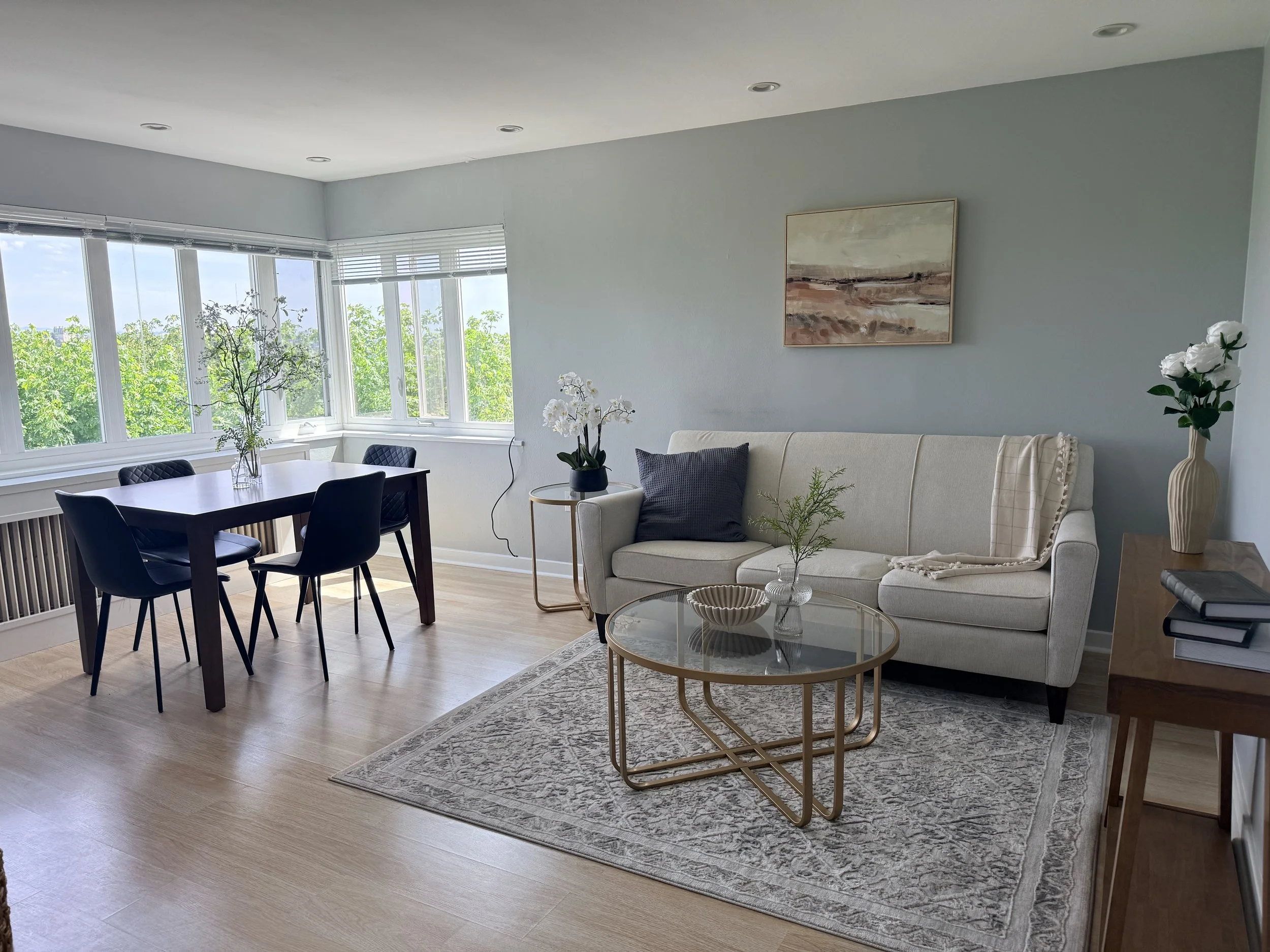 A living room with a dining table, black chairs, a beige sectional sofa, round coffee table, and large windows showing greenery outside. Decor includes orchids, wall art, and a patterned rug.